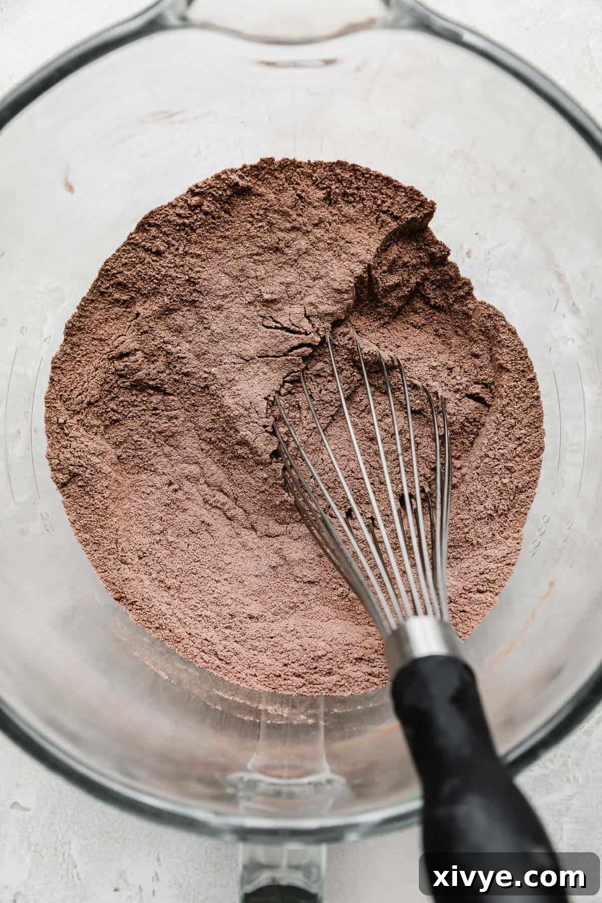 A glass bowl containing a mixture of dry ingredients like flour, sugar, and cocoa powder, with a wire whisk resting inside.