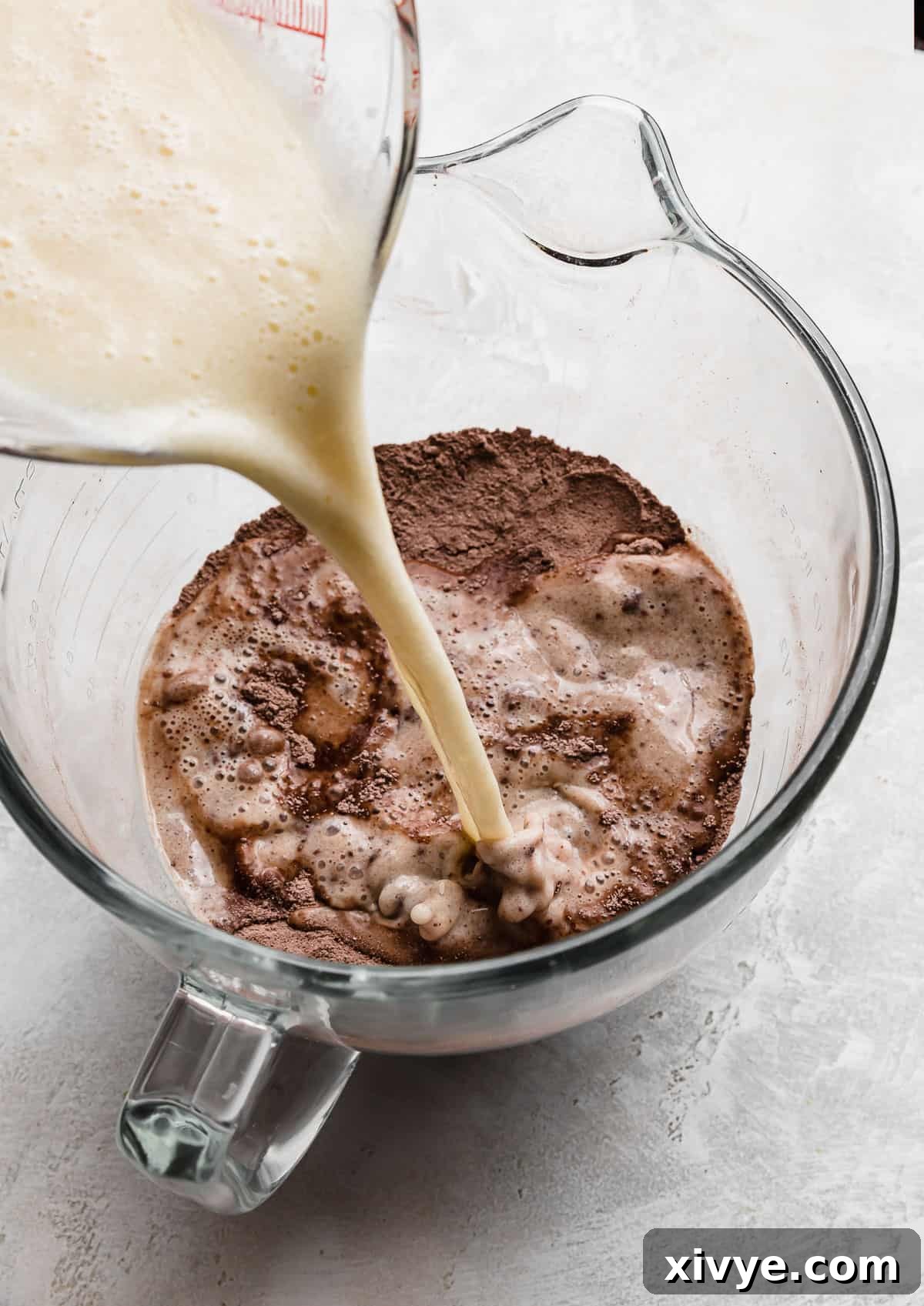 A stream of light yellow liquid wet ingredients being poured into a glass bowl already containing the brown dry ingredients, creating the rich batter for a German Chocolate Cake.