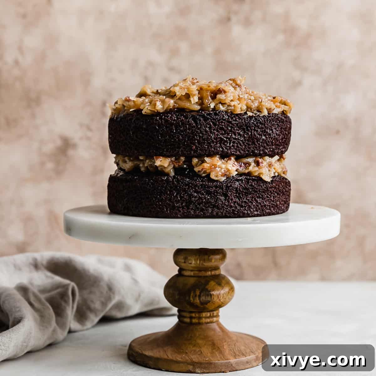 A two-layer Small German Chocolate Cake assembled on a white marble cake stand, showcasing the vibrant coconut pecan frosting between the layers, against a soft brown background.
