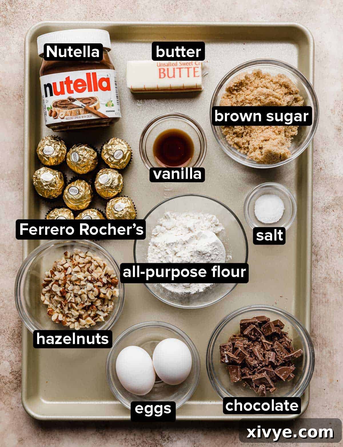 Ferrero Rocher Brownie ingredients in glass bowls on a bronze baking sheet on a light brown textured background.