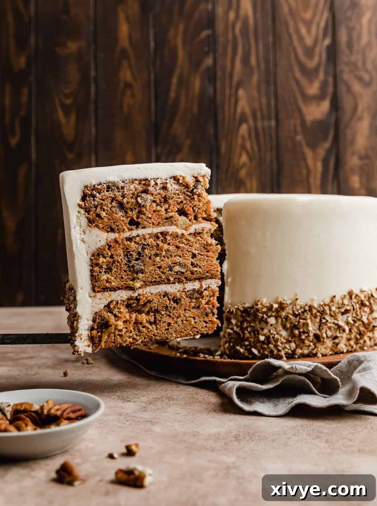 Sunshine Pineapple Pecan Carrot Cake 2 A slice of Carrot Cake with Pineapple and Pecans against a brown background with the cream cheese frosted carrot cake in the background.