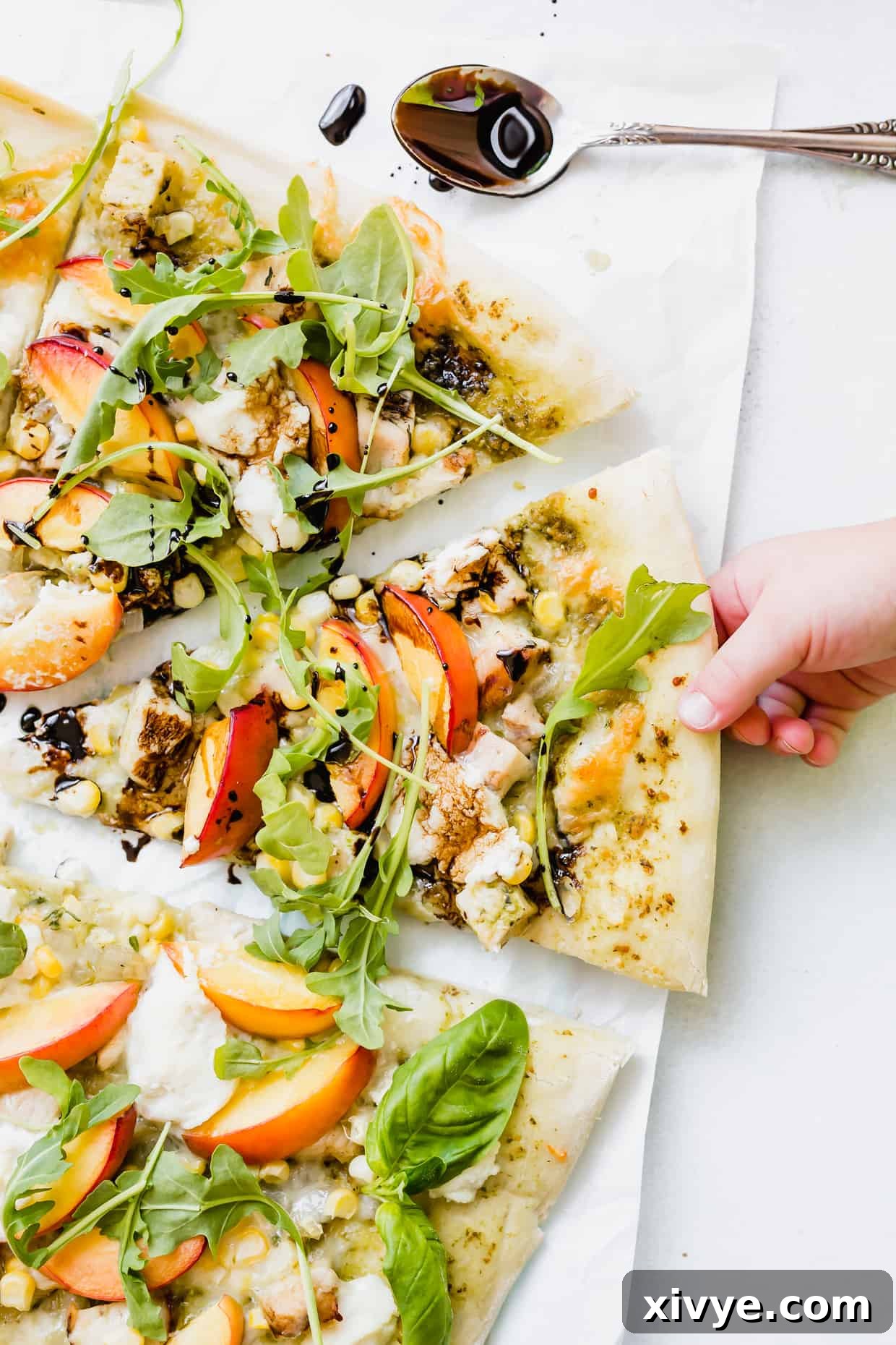 Peppery Rocket Pesto Pizza 2 A child's hand grabbing a slice of pizza that is topped with chicken, nectarines, arugula, and a balsamic drizzle.