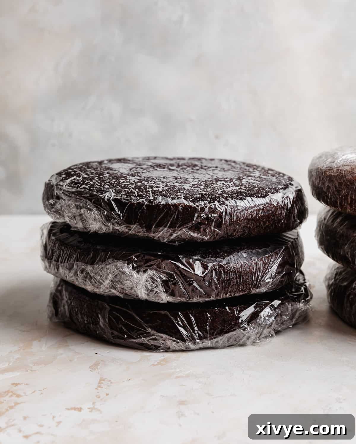 Three baked chocolate cake layers individually wrapped in plastic wrap, stacked on top of each other. 