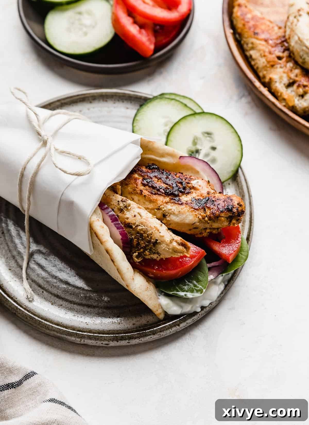 A chicken gyro made with chicken tenders wrapped in white parchment paper, on a grey plate.