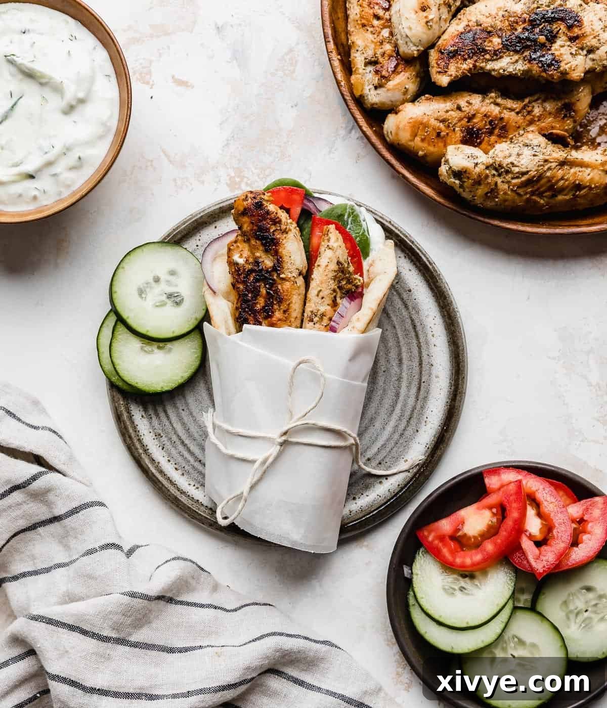 A Greek Chicken Gyro topped with tomatoes, chicken tenders, and wrapped in white parchment on a white background.