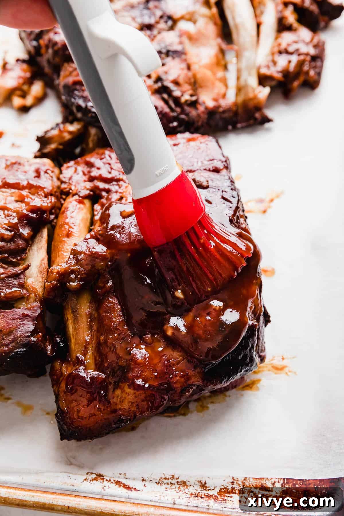 A red pastry brush gracefully spreading a dark, rich Coca Cola rib sauce over perfectly cooked pork ribs, preparing them for the final caramelization.