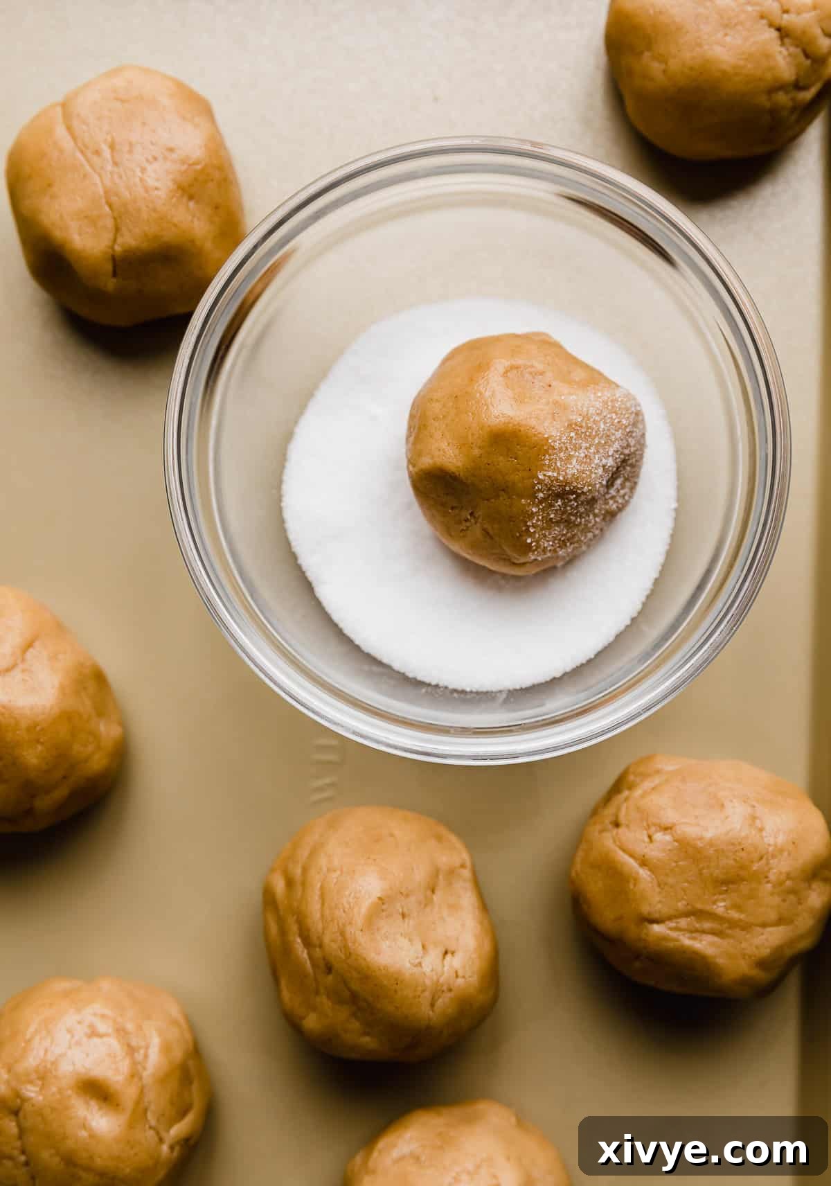 Crumbl's Iconic Peanut Butter Perfection 5 Light brown peanut butter cookies rolled in balls with one ball in a glass bowl that's filled with white sugar.