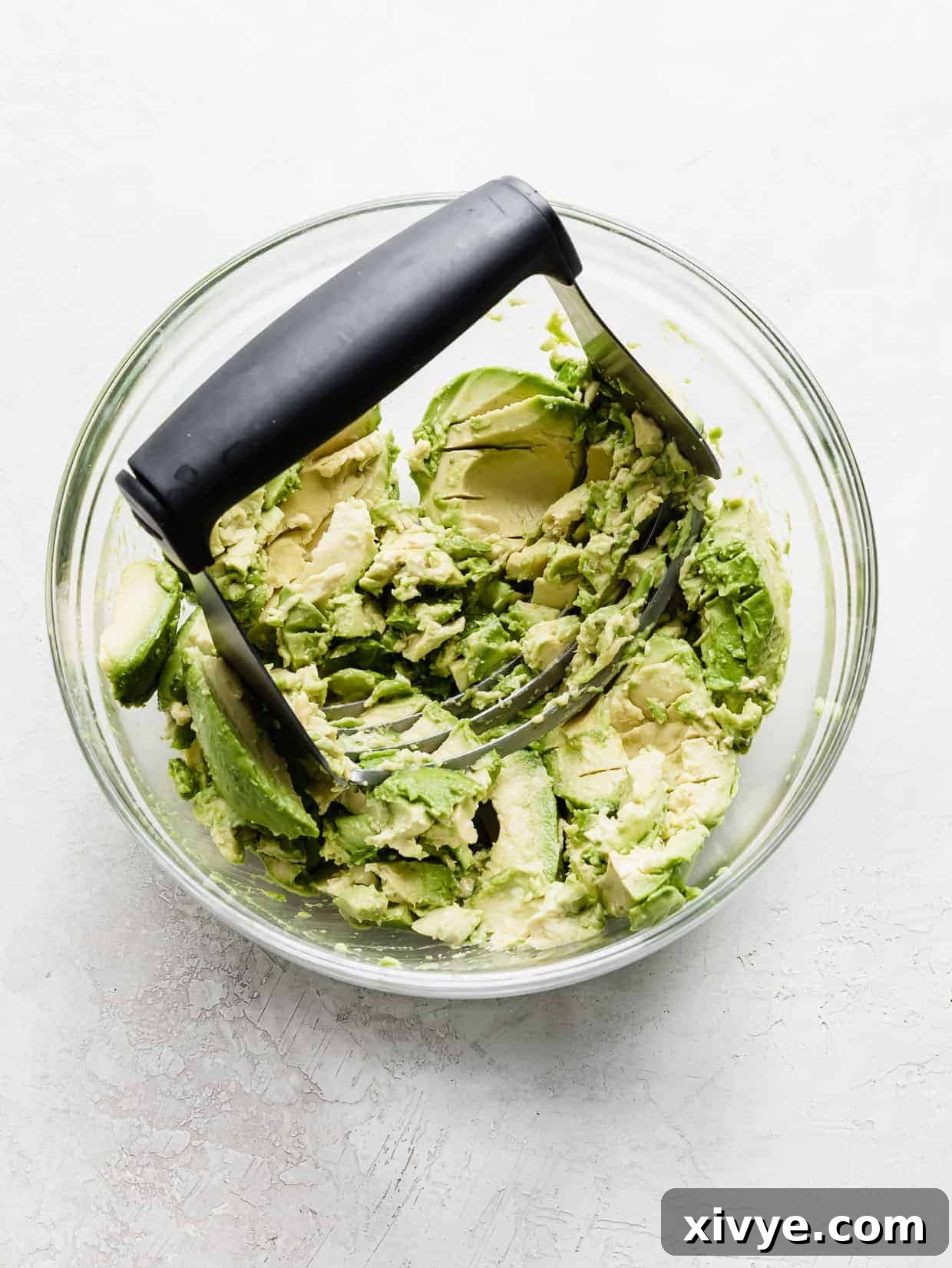 Fresh avocados in a glass bowl with a pastry blender smashing them. 