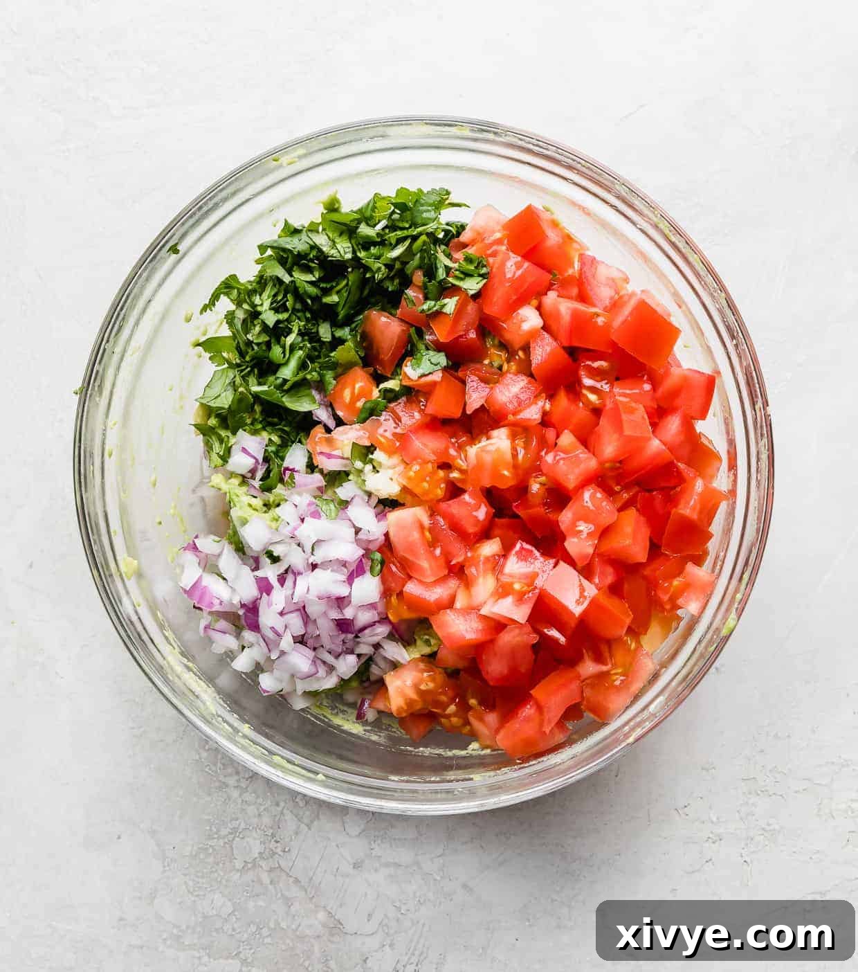 A glass bowl with avocado, fresh tomato, onion, and cilantro in it. 