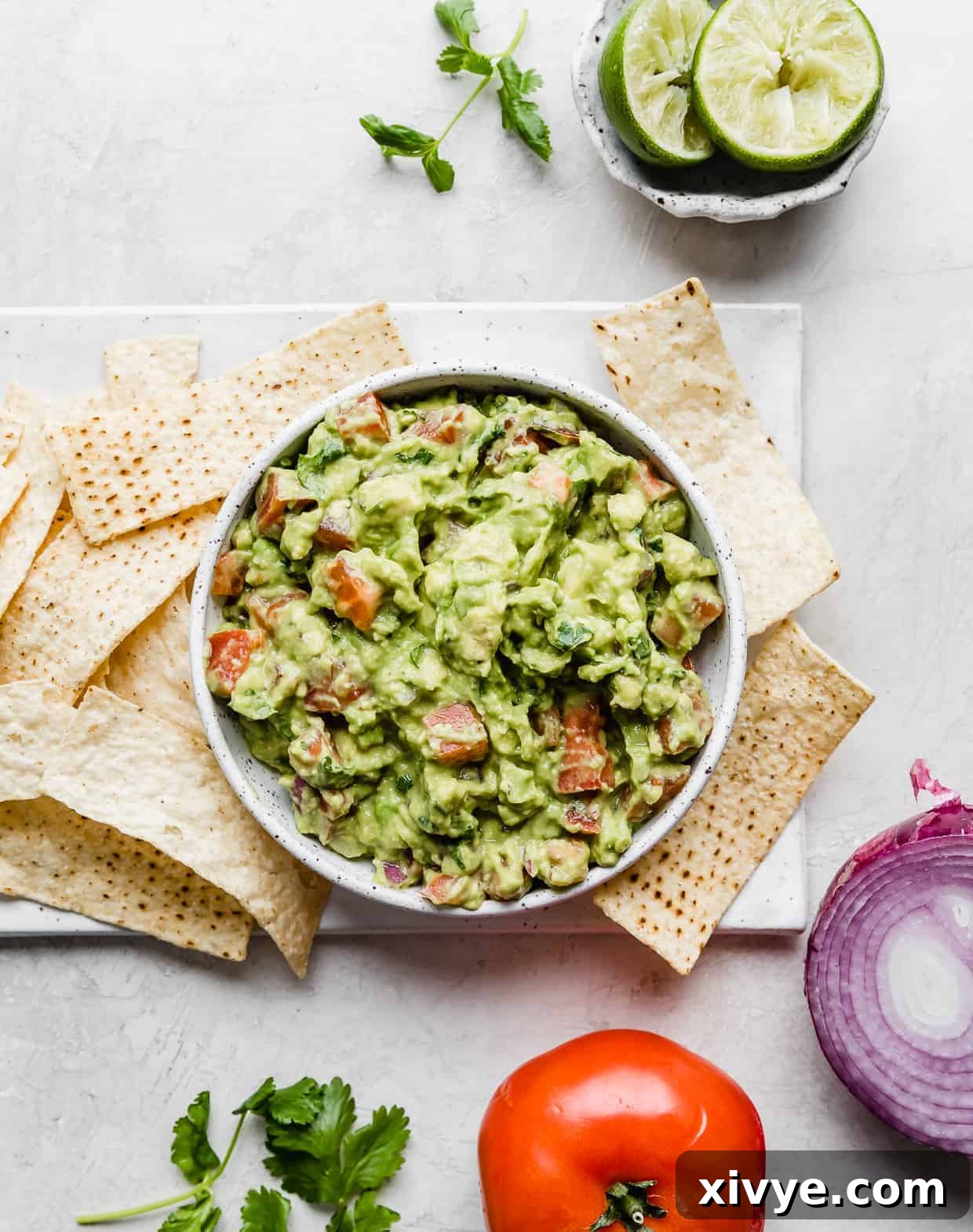 Overhead photo of the best guacamole in a bowl surrounded by a red onion, tomato, and limes.