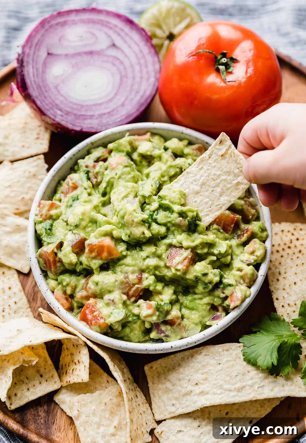 A small hand dipping a tortilla chip into a bowl of fresh guacamole with diced tomatoes in it.