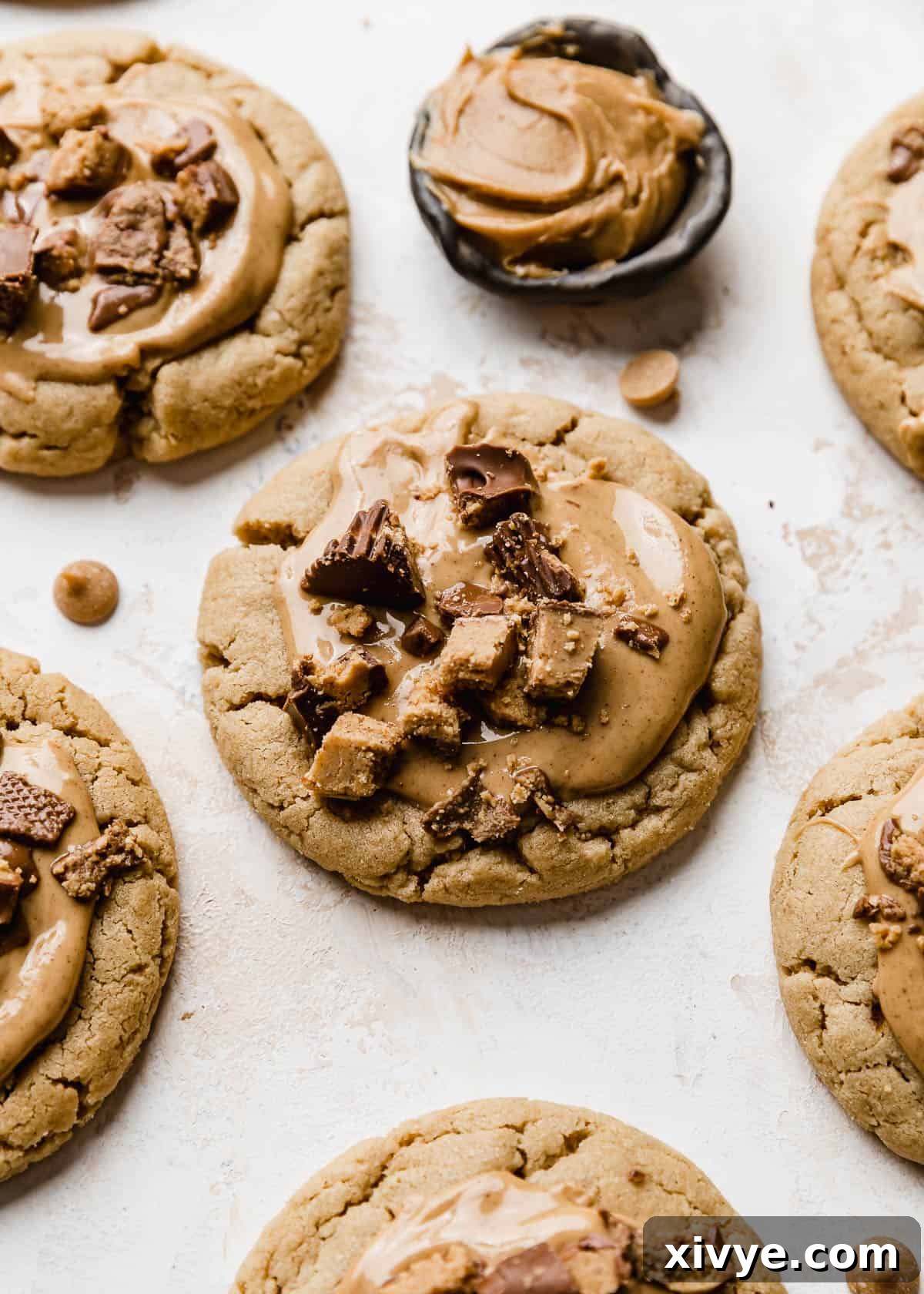 A delicious Crumbl Peanut Butter Cup Cookie, generously topped with a smooth layer of melted peanut butter and adorned with chopped Reese's peanut butter cups, resting on a white surface.