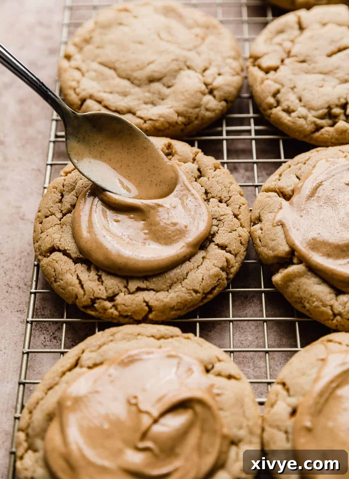 A hand using a spoon to gracefully spread a smooth layer of melted peanut butter topping over a warm peanut butter cookie.