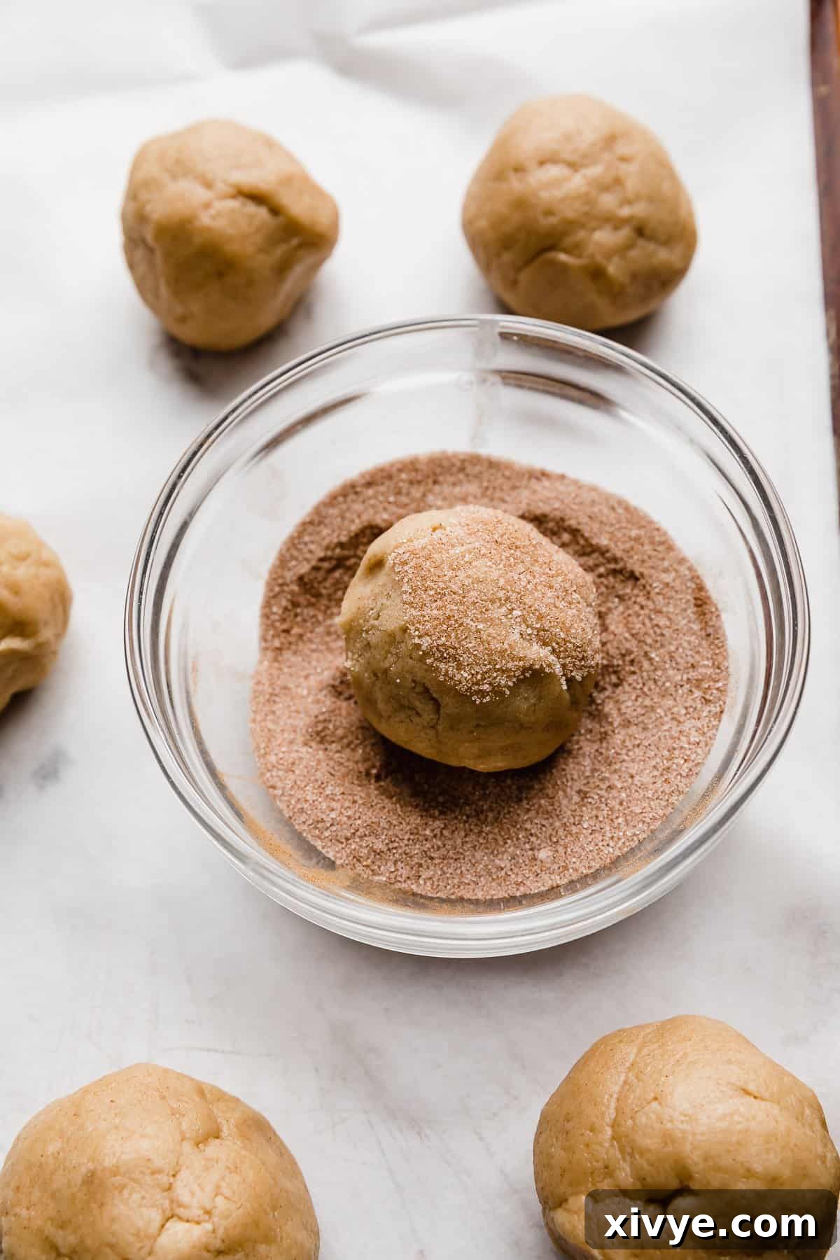 Homemade Crumbl Churro Cookie Perfection 5 A perfectly spherical ball of churro cookie dough is captured mid-roll in a shallow bowl brimming with cinnamon sugar, ready for baking.