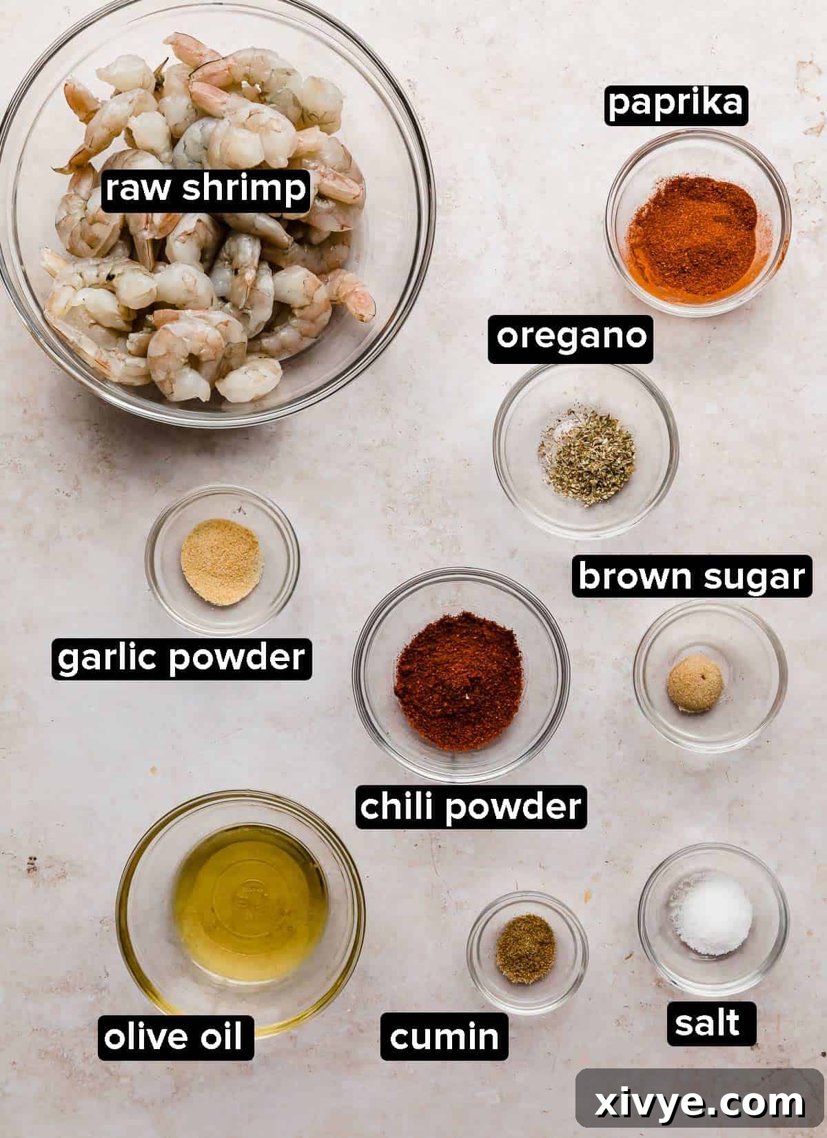 Various ingredients for Blackened Shrimp, including spices and fresh shrimp, neatly portioned into glass bowls on a textured beige background.