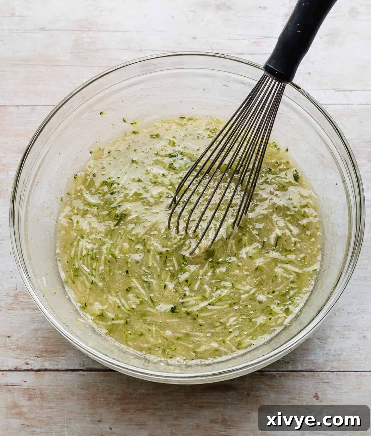 Shredded zucchini in a glass bowl with a whisk.