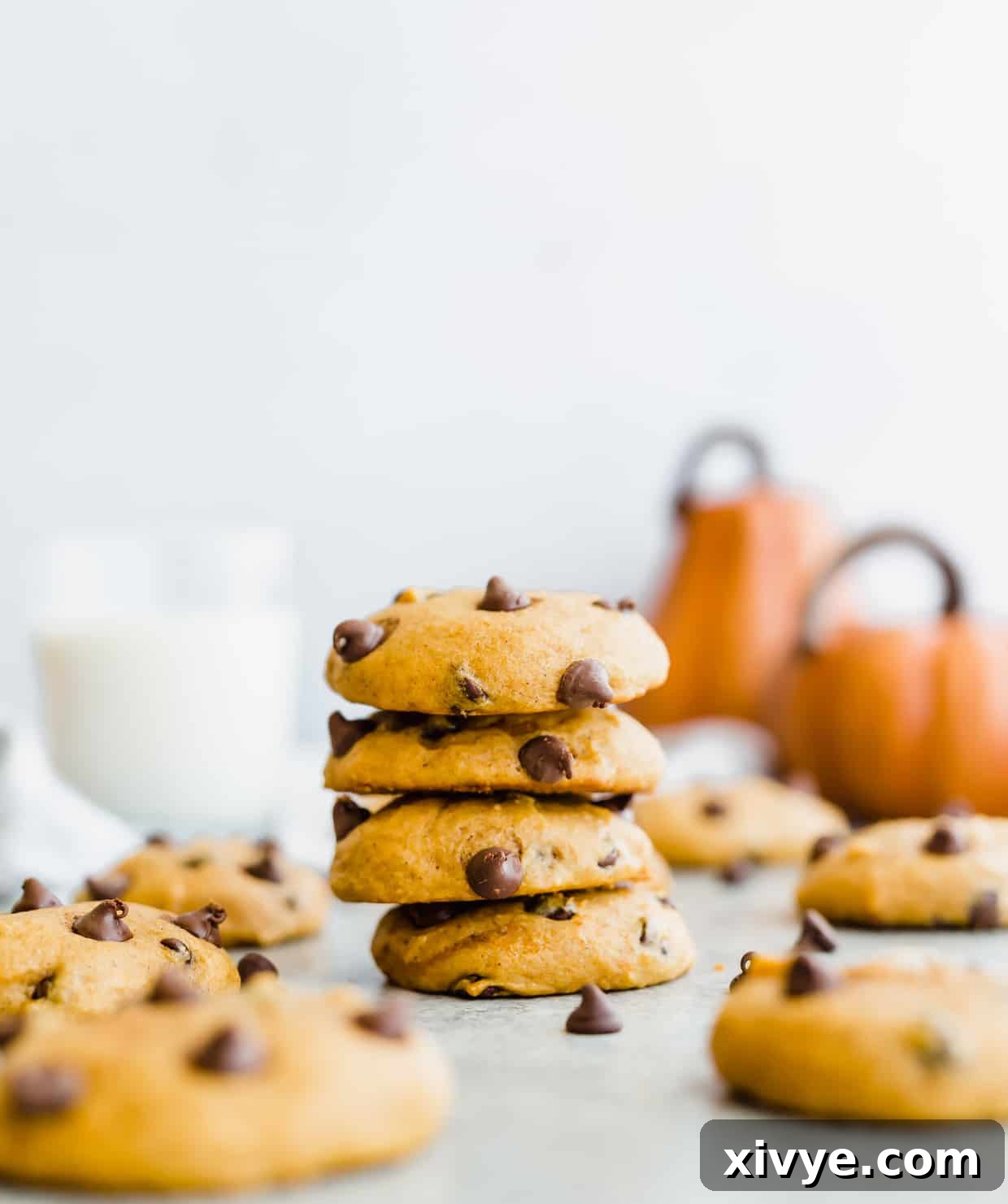A stack of 4 perfectly baked, soft pumpkin chocolate chip cookies with visible chocolate chips, beautifully arranged with two small decorative pumpkins in a warm, inviting background.