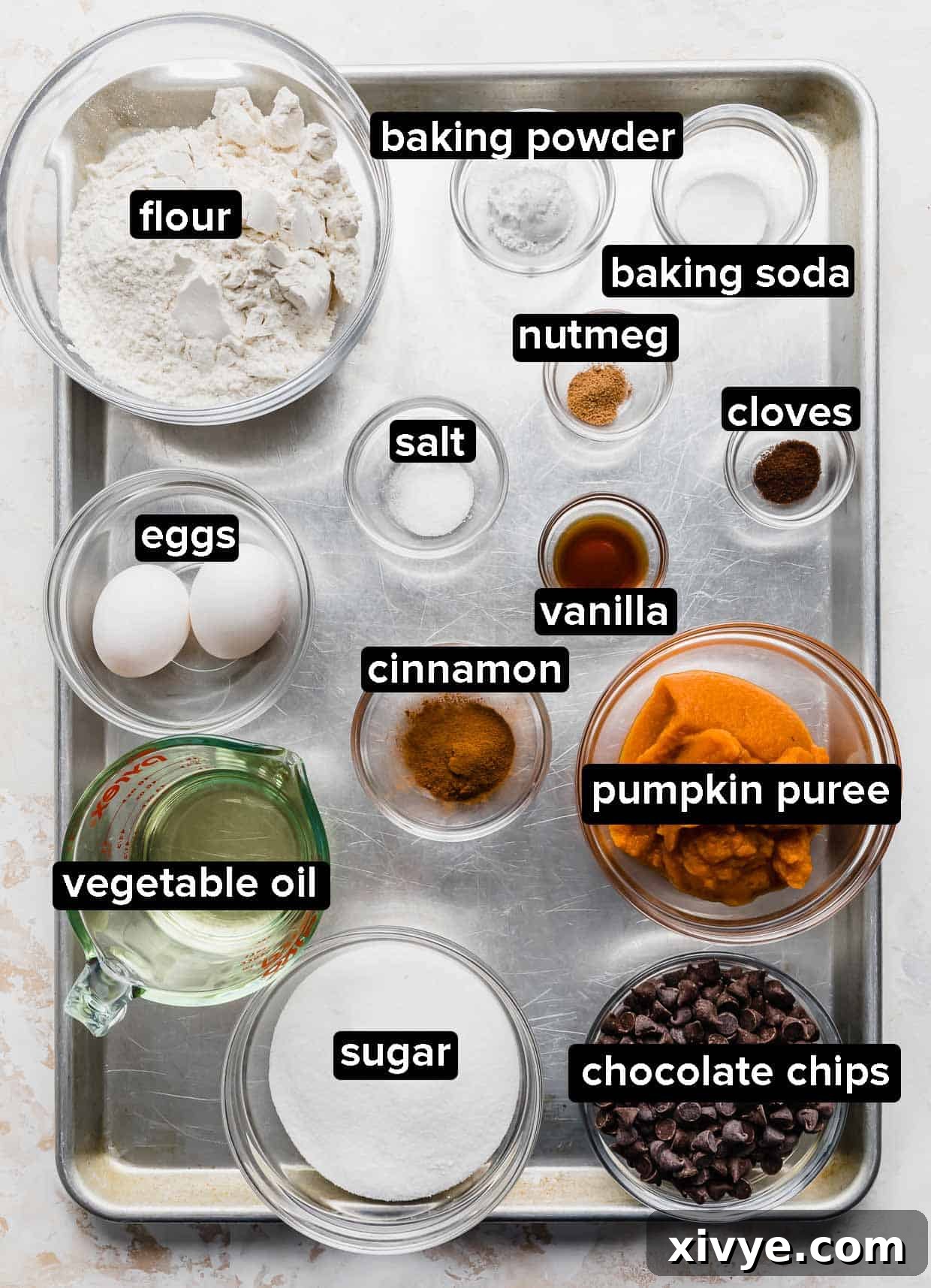 All the fresh, high-quality ingredients for making Pumpkin Chocolate Chip Cookies, including flour, spices, pumpkin puree, chocolate chips, eggs, and vegetable oil, neatly arranged on a silver baking sheet.