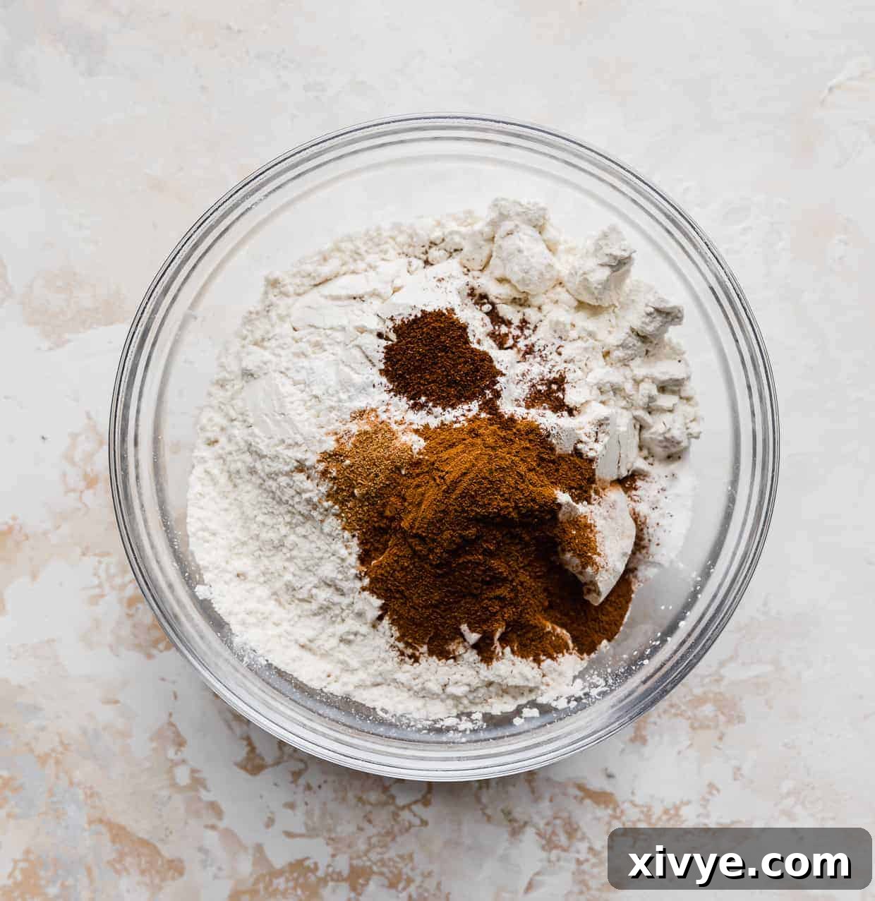 Dry ingredients for Pumpkin Chocolate Chip Cookies, including flour, baking powder, baking soda, salt, cinnamon, nutmeg, and cloves, measured out in a clear glass bowl on a textured cream background.
