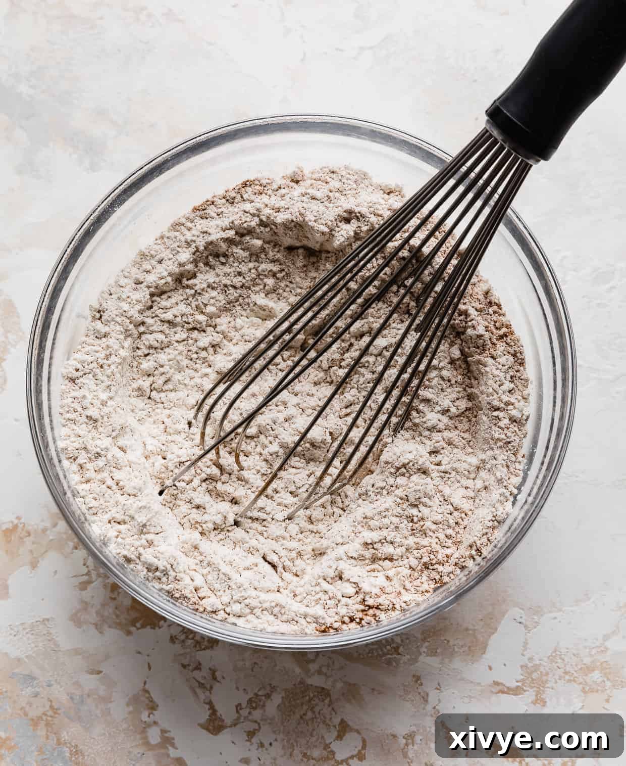 A clear glass bowl with a whisk stirring dry ingredients together for pumpkin chocolate chip cookies, creating a fine, uniform mixture.