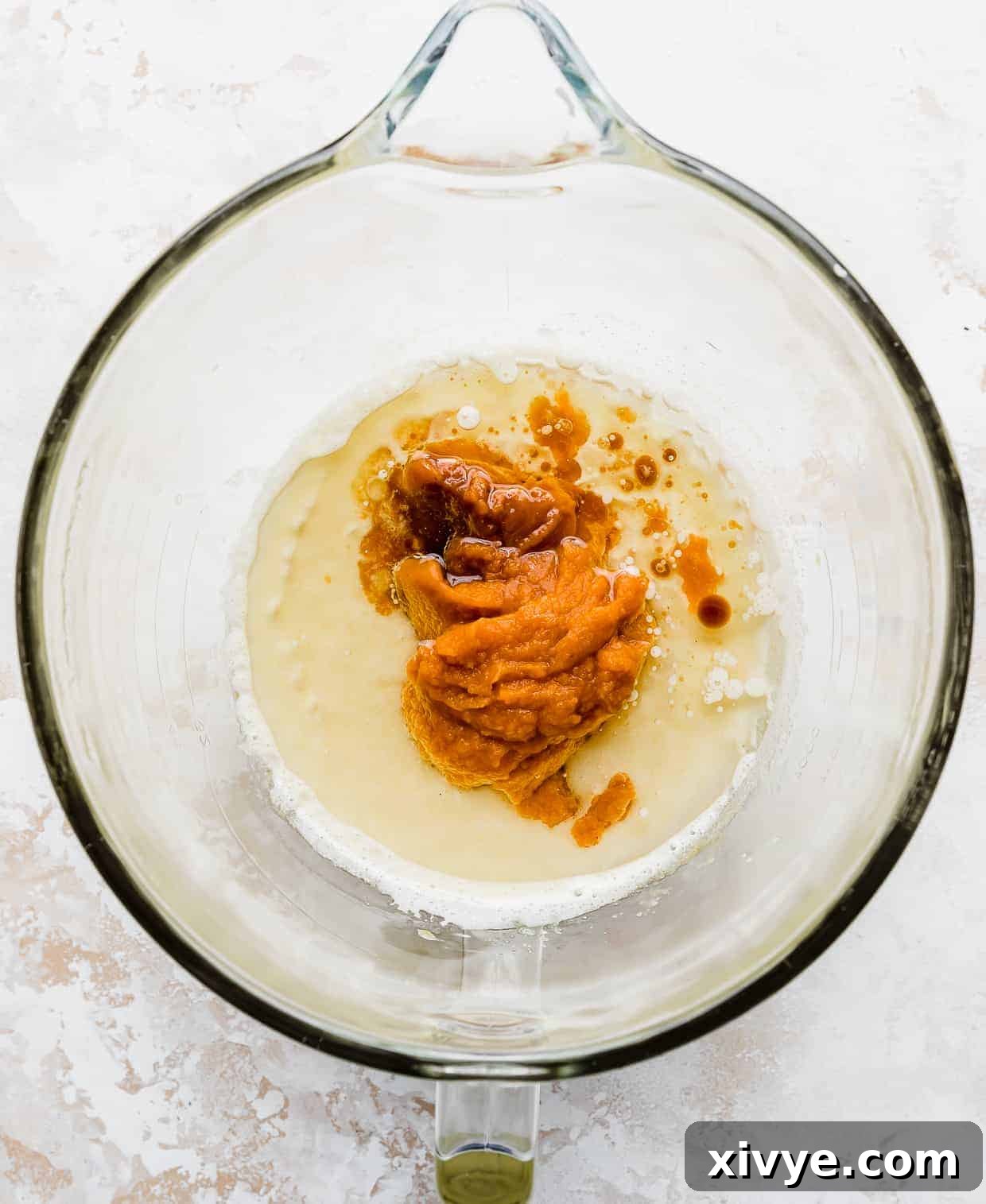 A scoop of vibrant orange pumpkin puree being added to the wet ingredients in a glass mixing bowl, for the Pumpkin Chocolate Chip Cookies recipe.