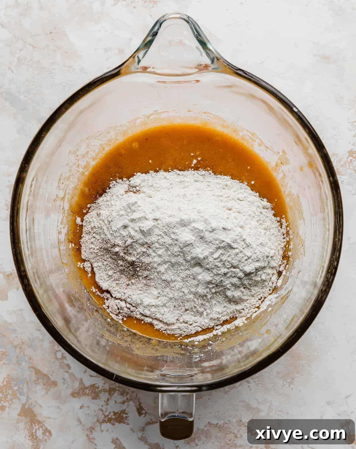 Flour and other dry ingredients dusted over an orange liquid mixture in a glass mixing bowl, awaiting combination into cookie dough.