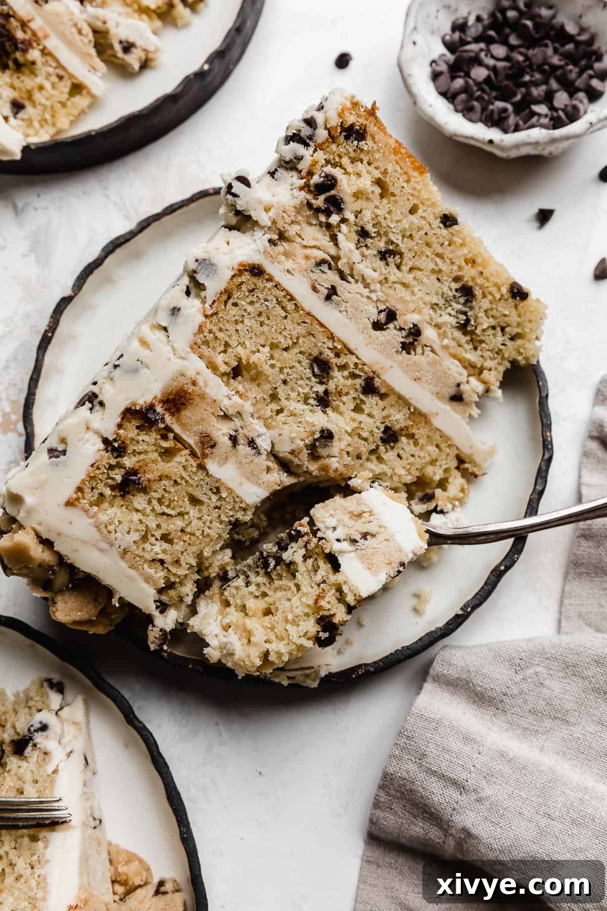 A Cookie Dough Cake slice on a black rimmed white plate with a fork cutting into the top portion of the cake.