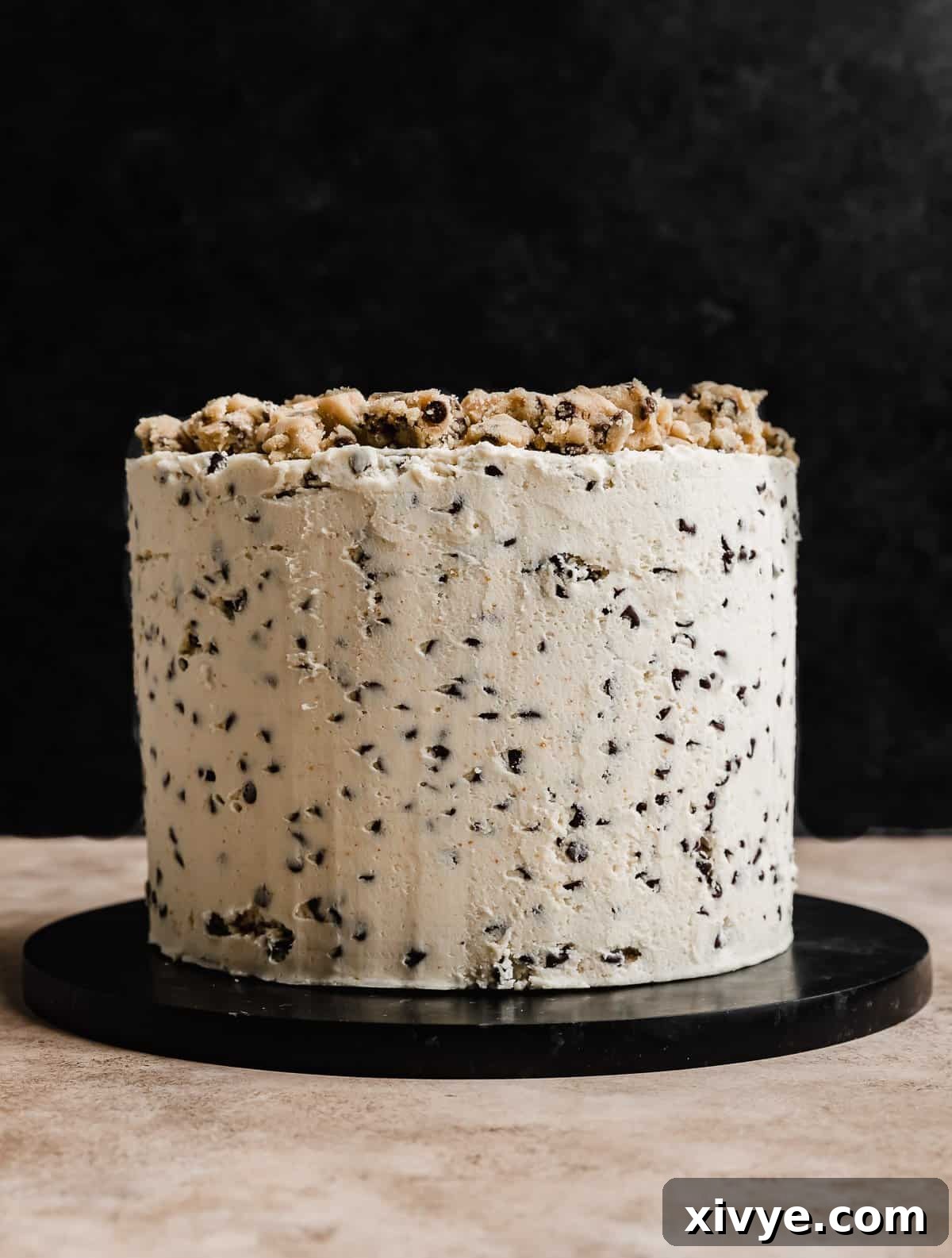 A three layered round Cookie Dough Cake frosted with white frosting that has mini chocolate chips in it, against a black background.