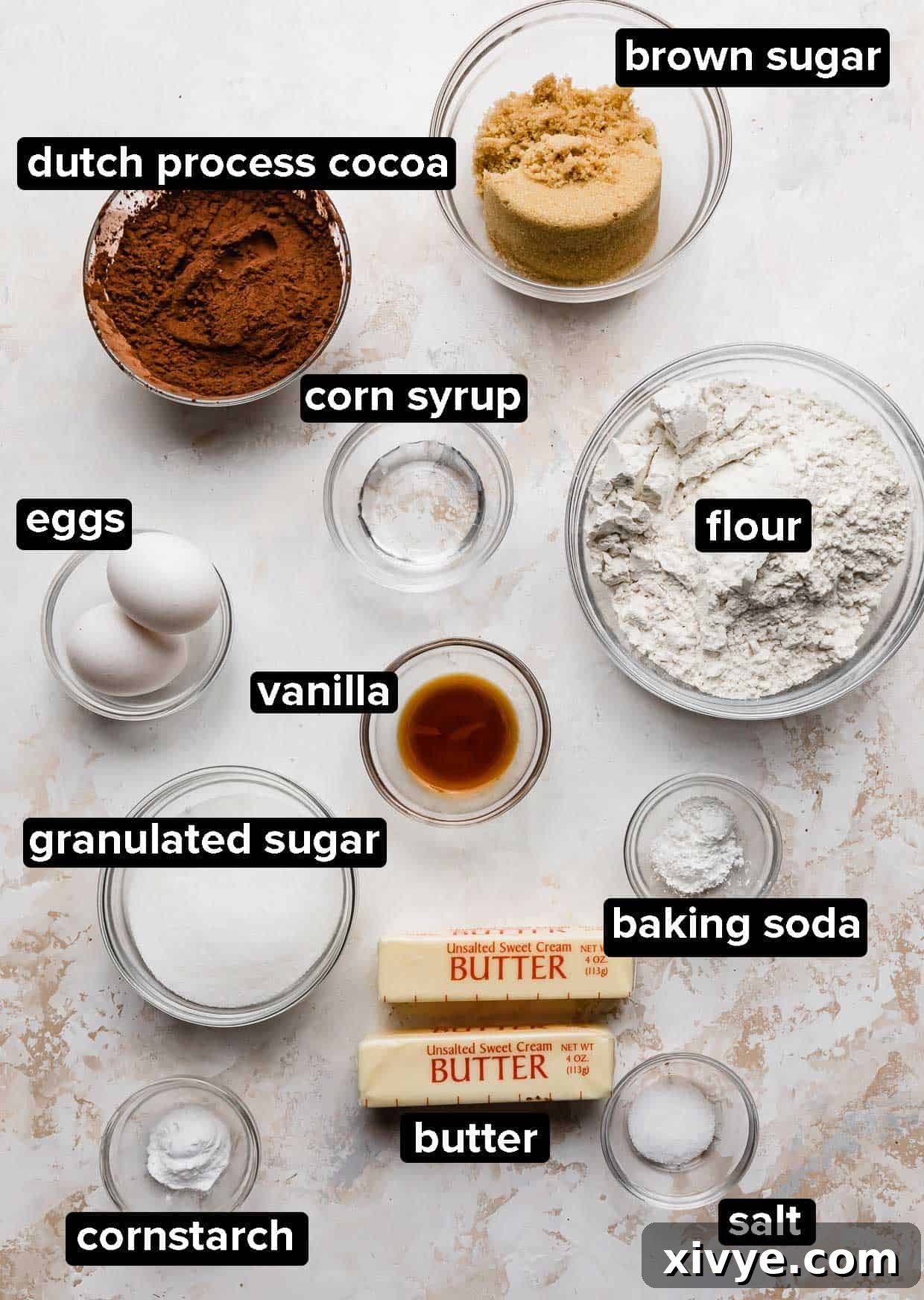 Decadent Mint Brownie Cookie Bliss 3 A selection of baking ingredients, including flour, cocoa powder, sugar, butter, and eggs, arranged neatly on a white textured background, ready for making cookies.