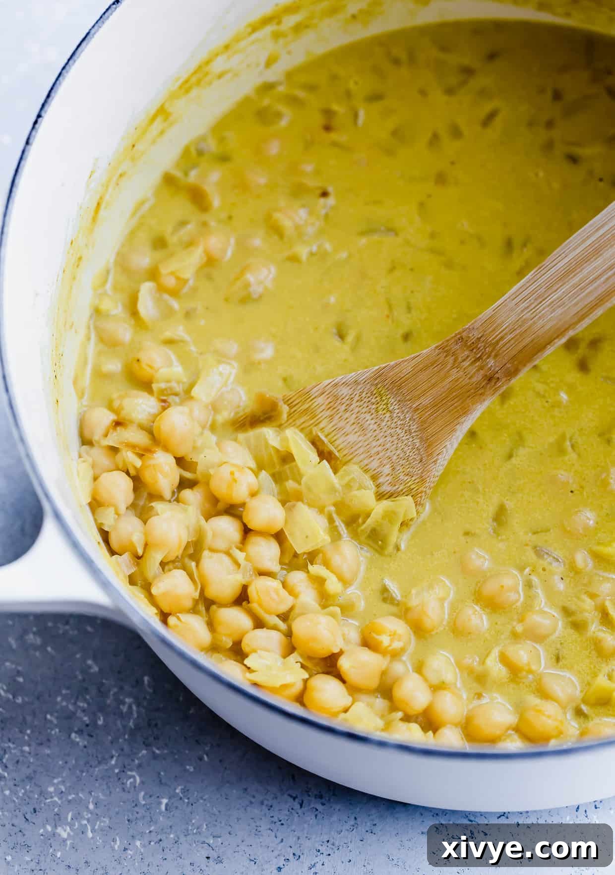 A close-up of a bubbling pot of chickpea curry, rich and creamy, with chickpeas visible and a wooden spoon lifting some out.