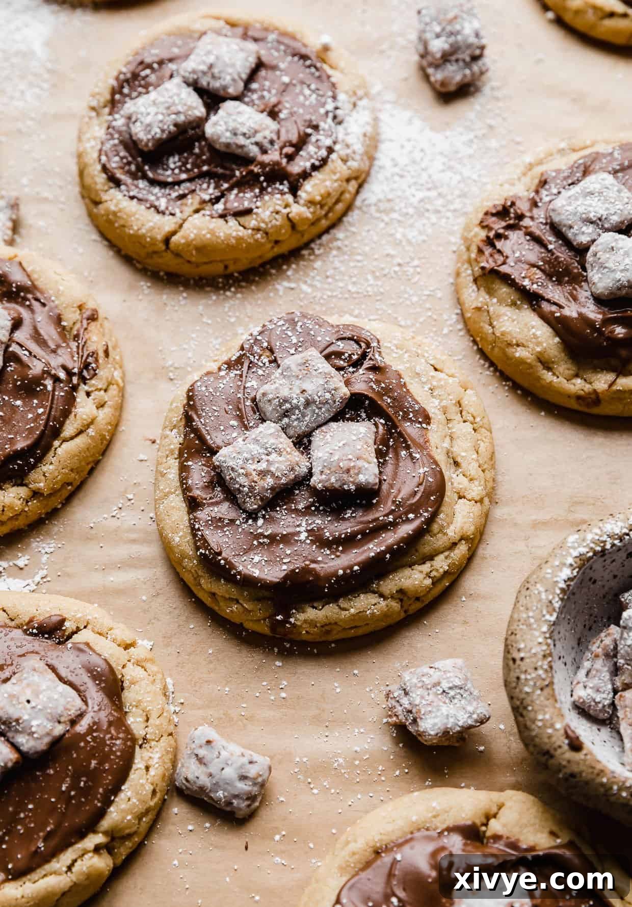 A peanut butter cookie topped with melted chocolate and three Muddy Buddy pieces, dusted with powdered sugar, sitting on a light brown parchment paper. The cookie is large and inviting.