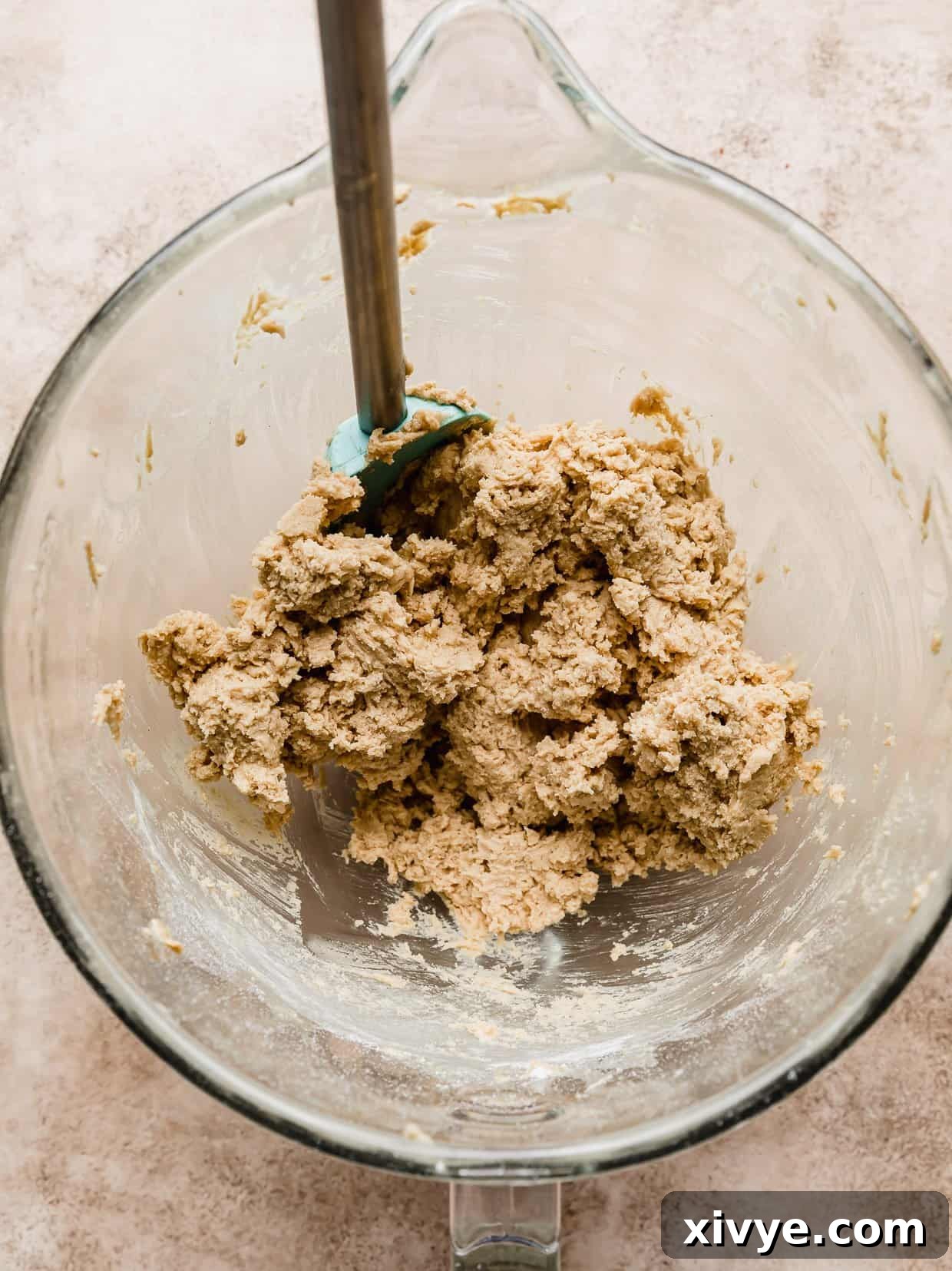 Peanut butter cookie dough in a glass mixing bowl on a light brown textured background, perfectly ready for portioning.