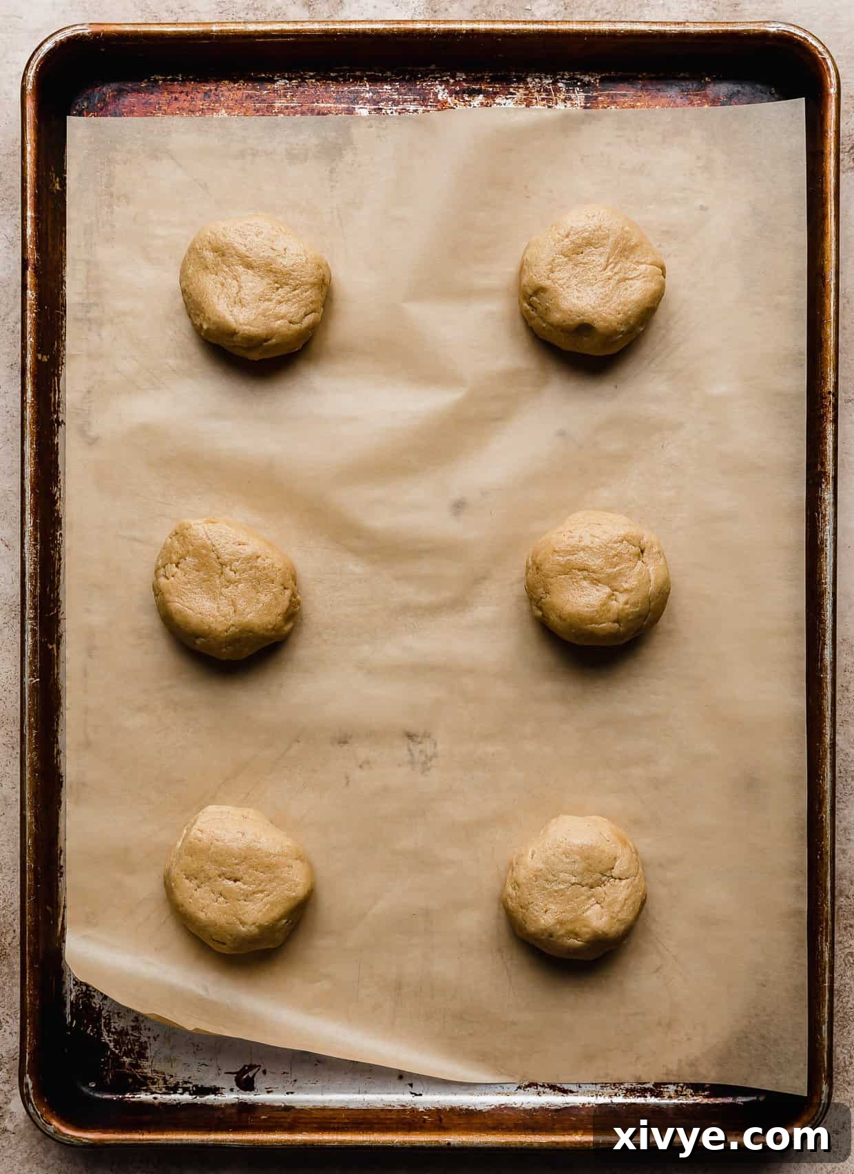 Six peanut butter cookie dough balls on a tan parchment lined baking sheet, flattened into thick disks, ready for baking to become muddy buddy crumbl cookies.