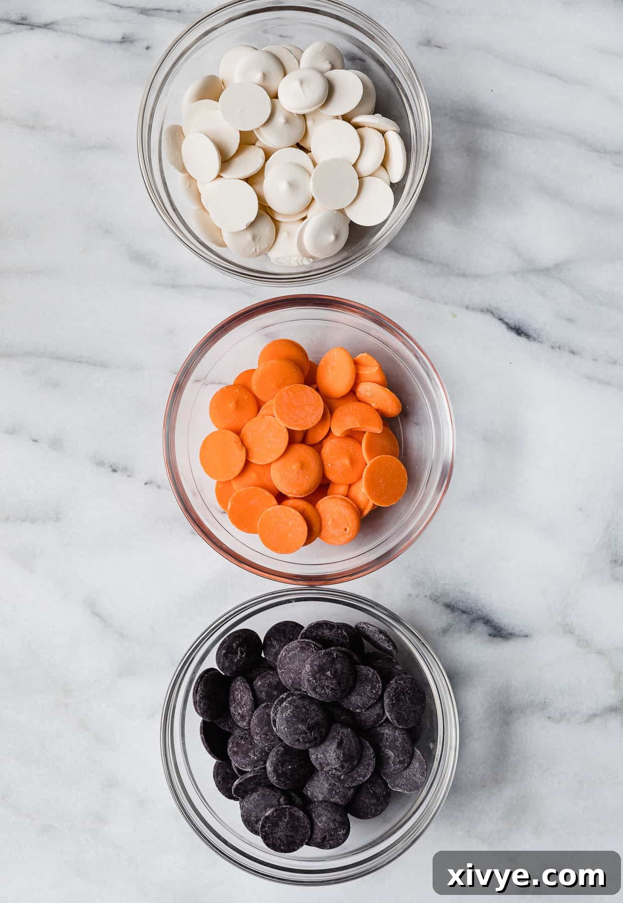 Three glass bowls filled with black, orange, and white melting chocolate disks on a white marble table.