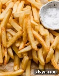 Golden French fries on a parchment paper with salt in a white bowl next to the fries.
