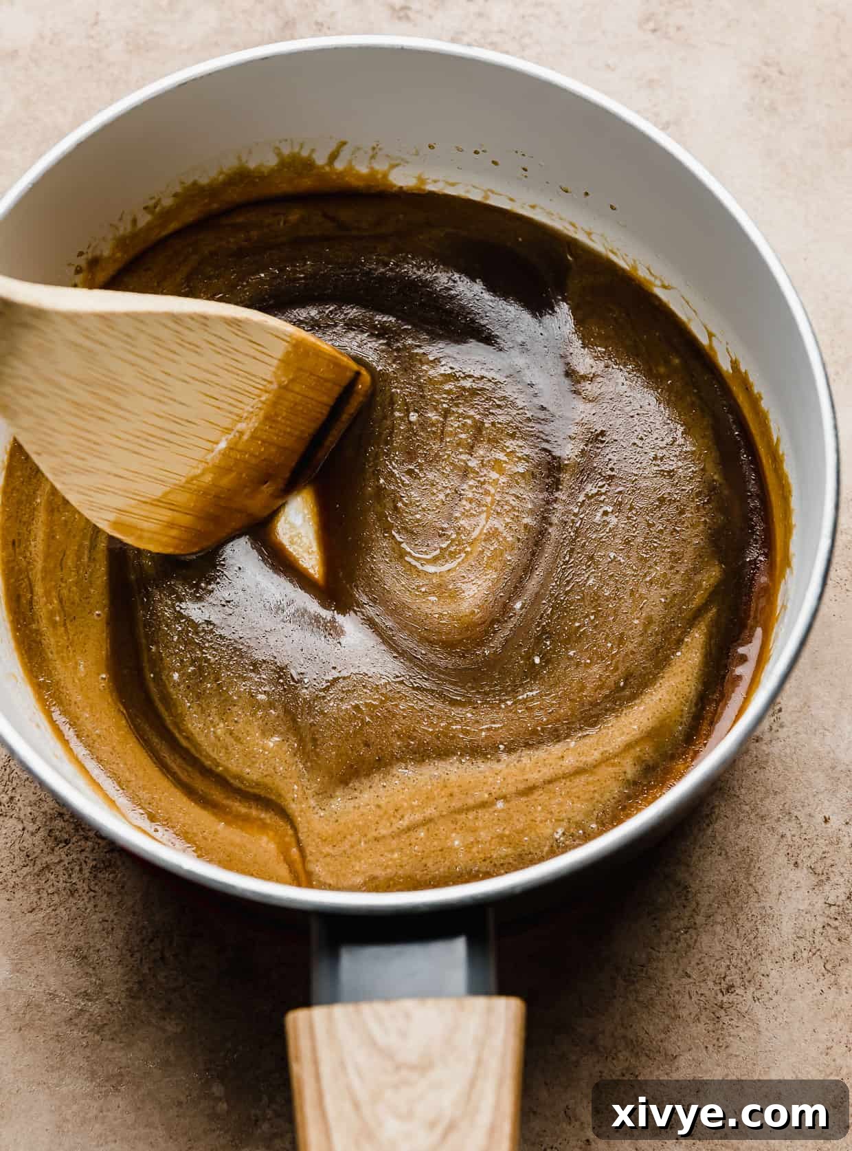 Salted Butterscotch Sauce simmering gently in a white pan, being stirred with a wooden spoon.