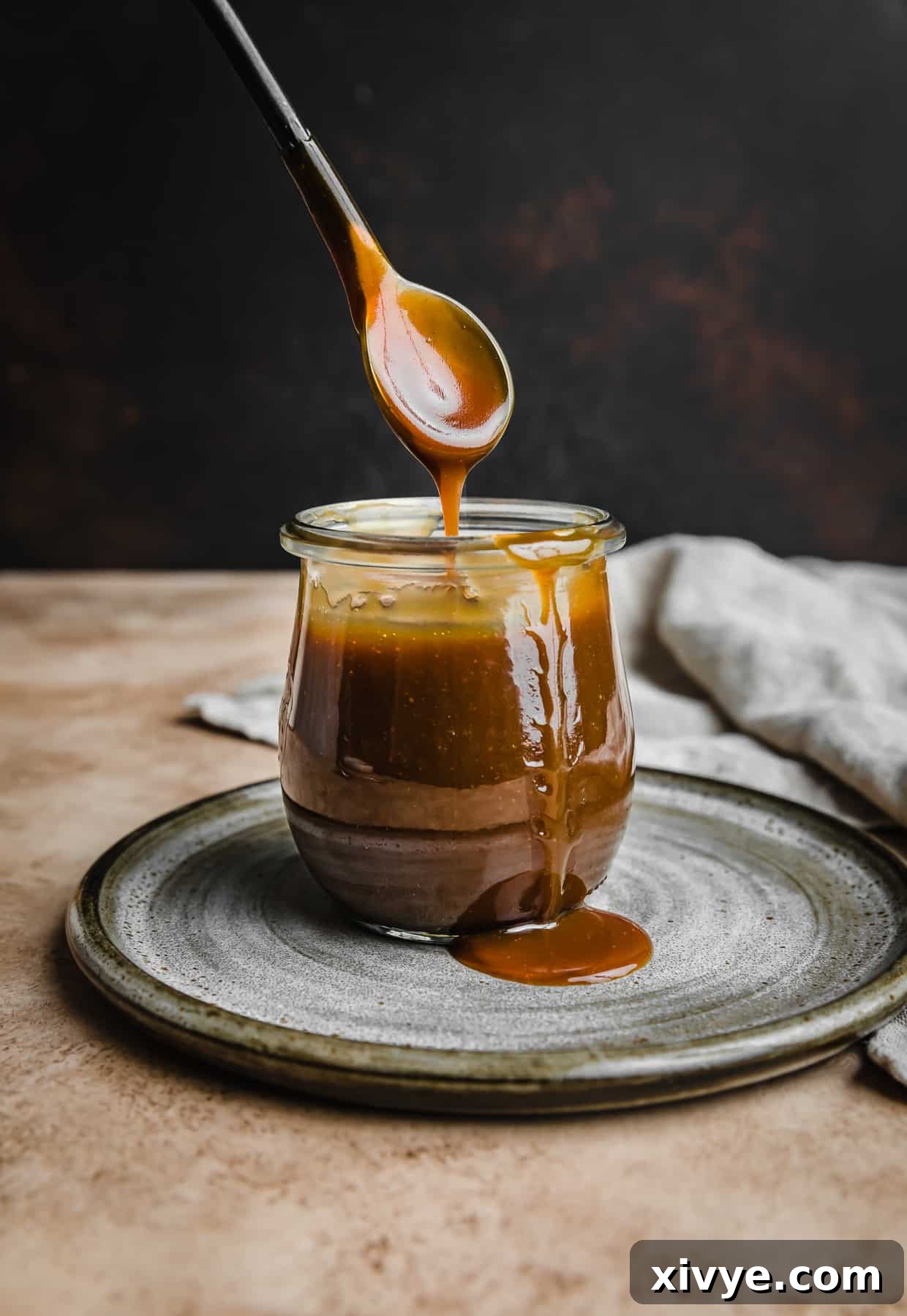 A spoon lifting out Salted Butterscotch topping from a small glass jar, against a dark brown background, showcasing its rich, thick texture.