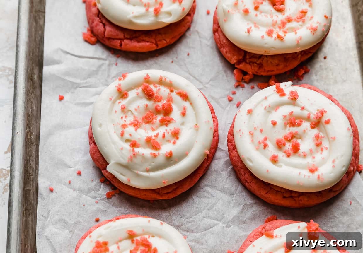 Crumbl Pink Velvet Cookies topped with frosting and pink velvet crumbs, on a white parchment lined baking sheet.