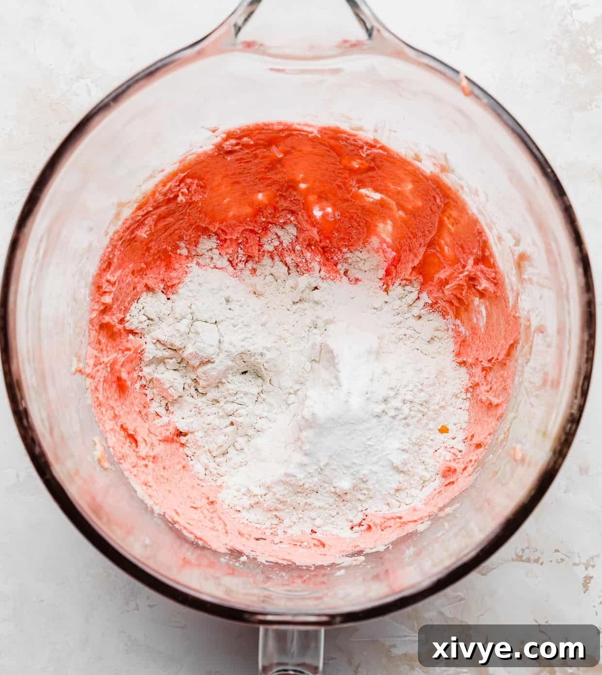 Flour overtop of pink cookie batter in a glass bowl.