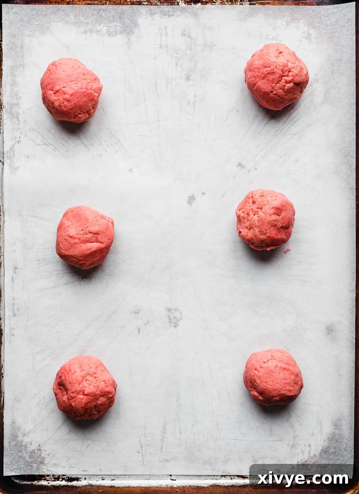 Six pink velvet cookie dough balls on a. white parchment lined baking sheet.