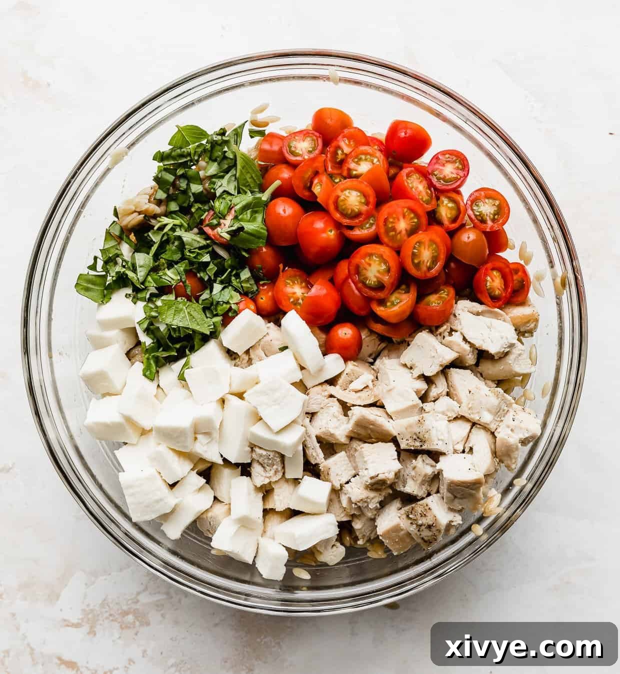 A large glass bowl filled with the vibrant components of Caprese Orzo Salad: mozzarella, tomatoes, basil, chopped chicken, and orzo pasta, before mixing.