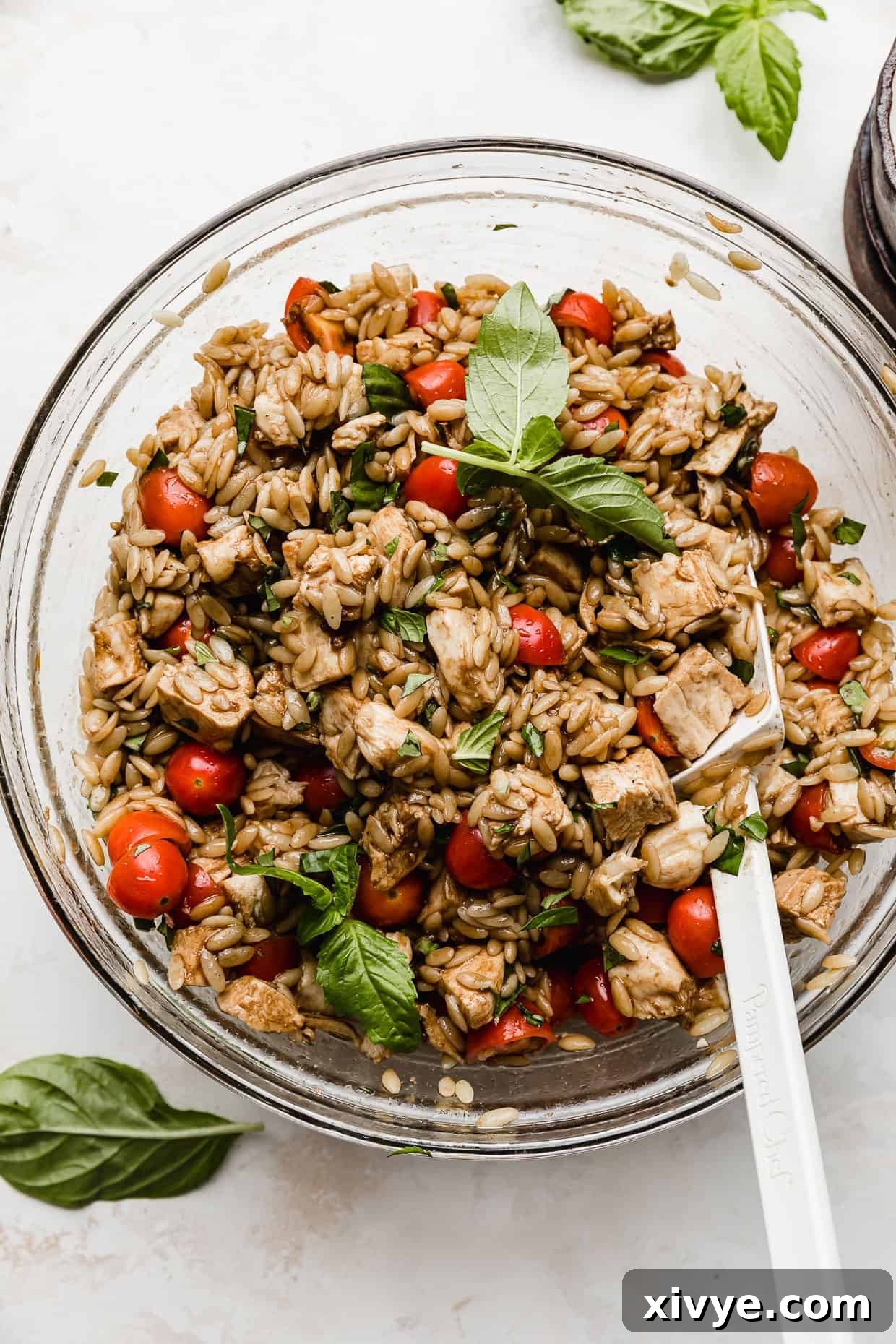A chef's hand gently stirring a Chicken Caprese Orzo Salad in a clear glass bowl with a white spatula, perfectly blending all the fresh ingredients.