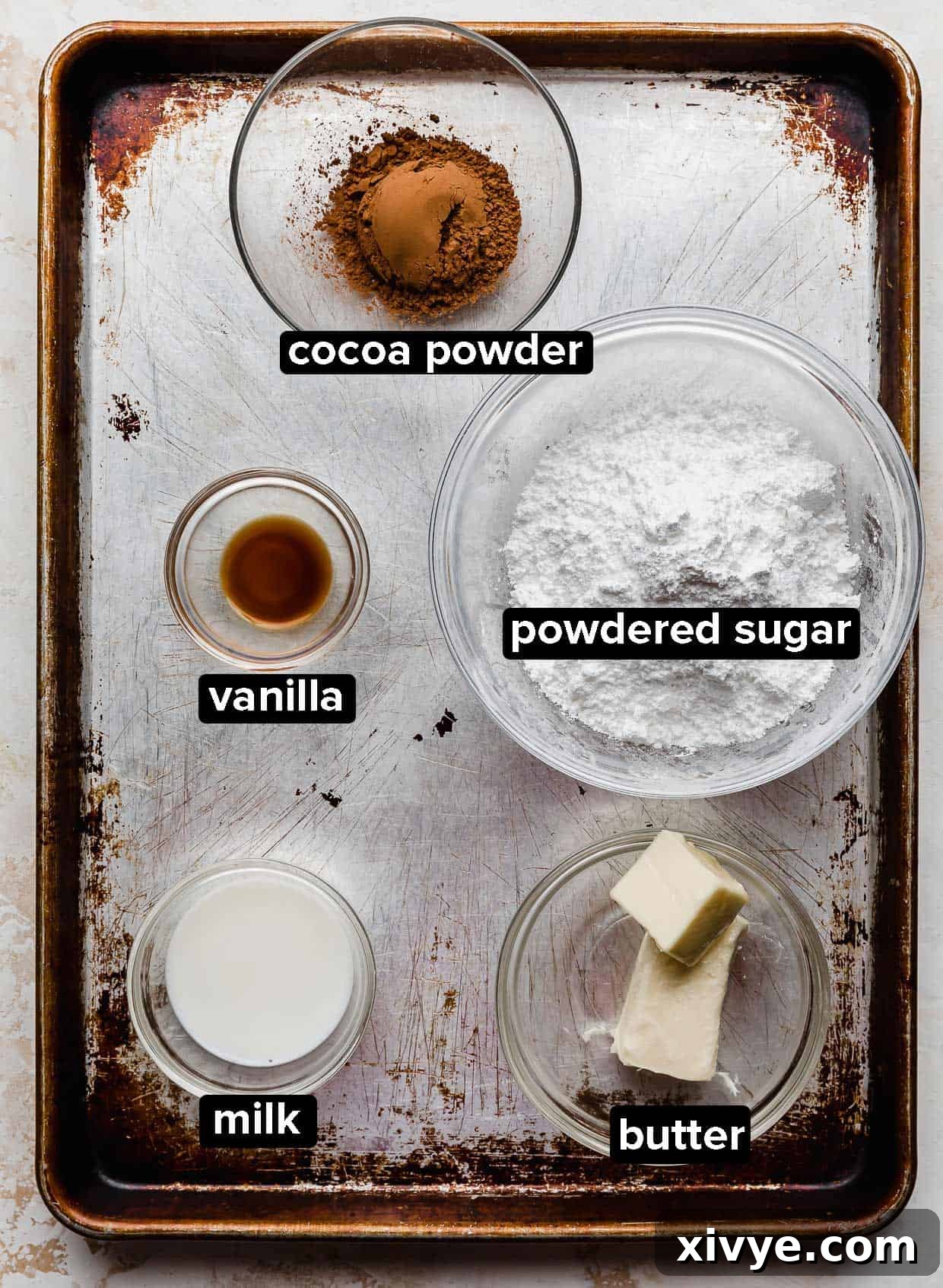 Ultimate Chocolate Chip Cookie Cake 4 Cookie cake chocolate frosting ingredients in glass bowls on a brown and gray worn baking sheet.