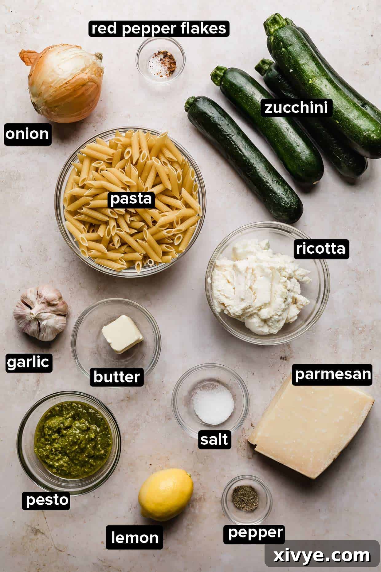 Ingredients laid out for making summer zucchini pasta with ricotta and basil, including fresh zucchini, basil, lemon, and cheese, on a cream background.