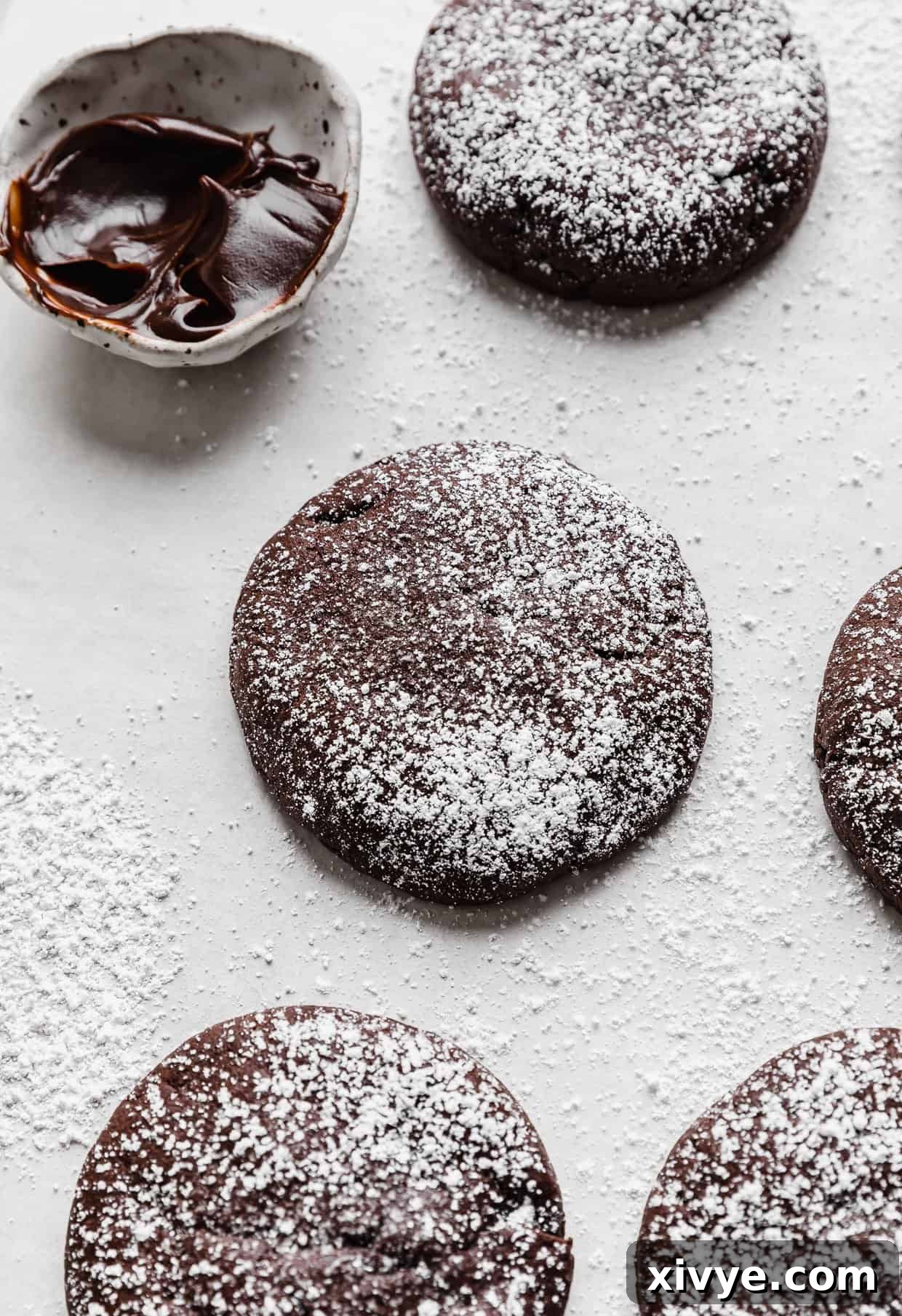 Powdered sugar dusted Crumbl Molten Lava Cookies on a white background with a small bowl of hot fudge in the background, ready to be served.