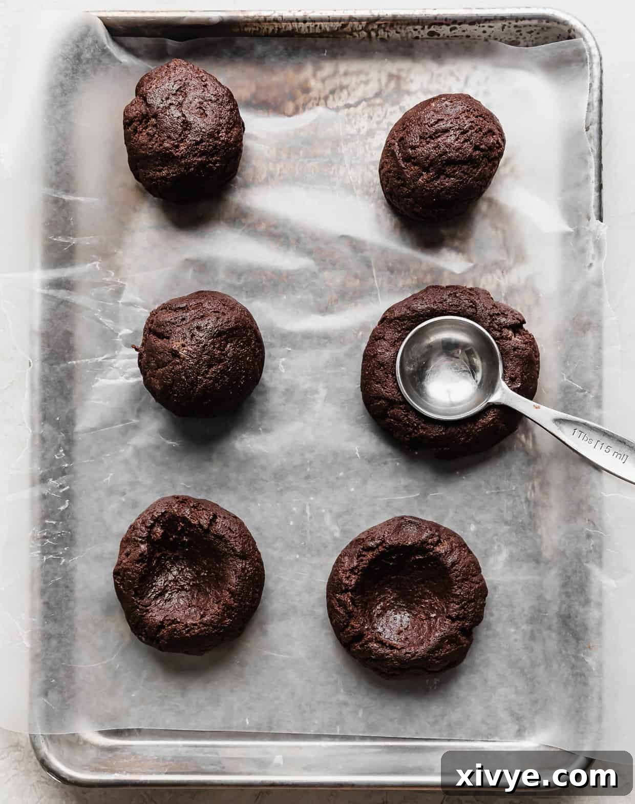 Six chocolate cookie dough balls with a metal tablespoon pressing into the center of one of the balls to create an indentation.