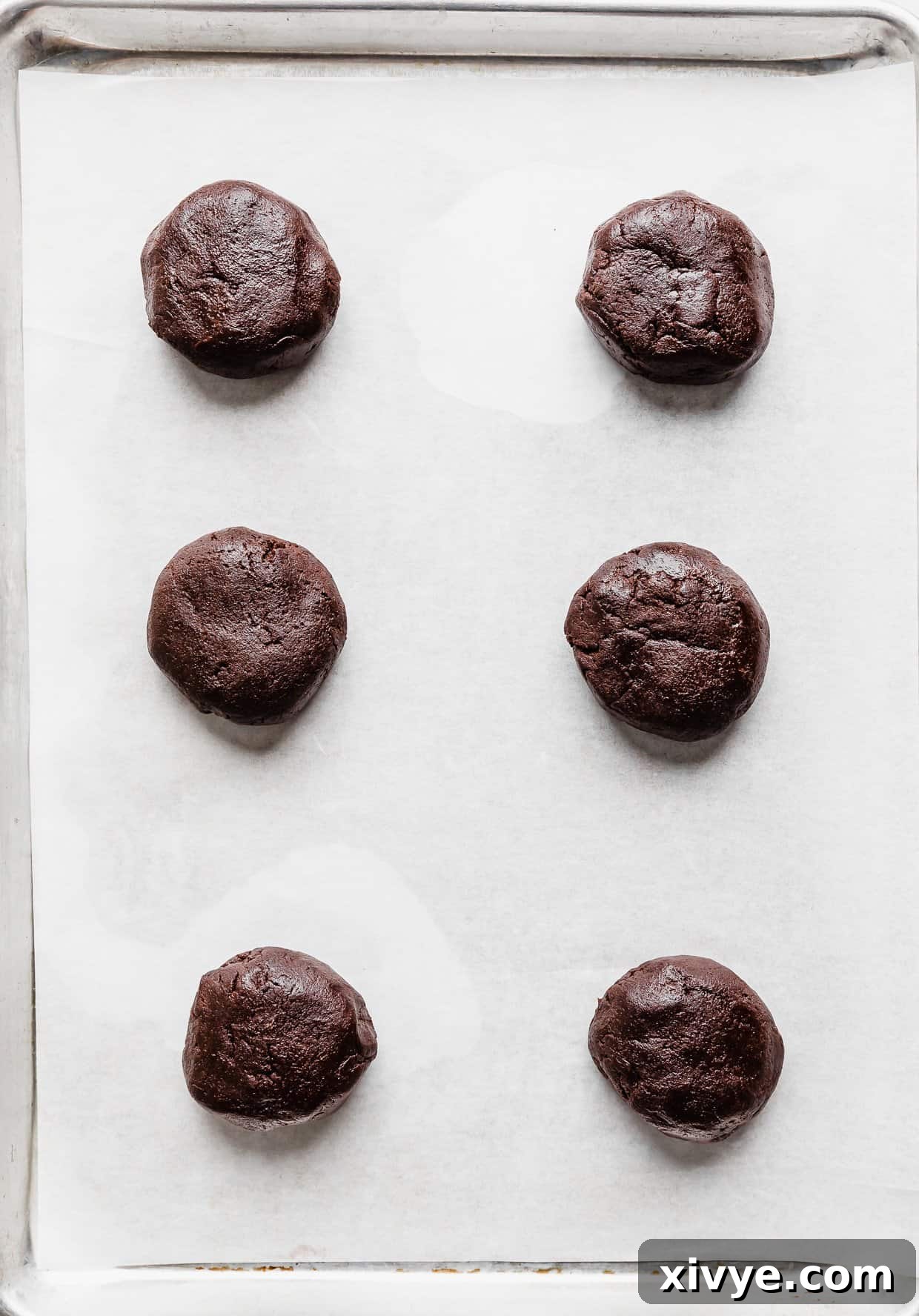 Six Crumbl Molten Lava Cookie dough balls, pressed into thick disks, on a white parchment paper lined baking sheet, ready for the oven.