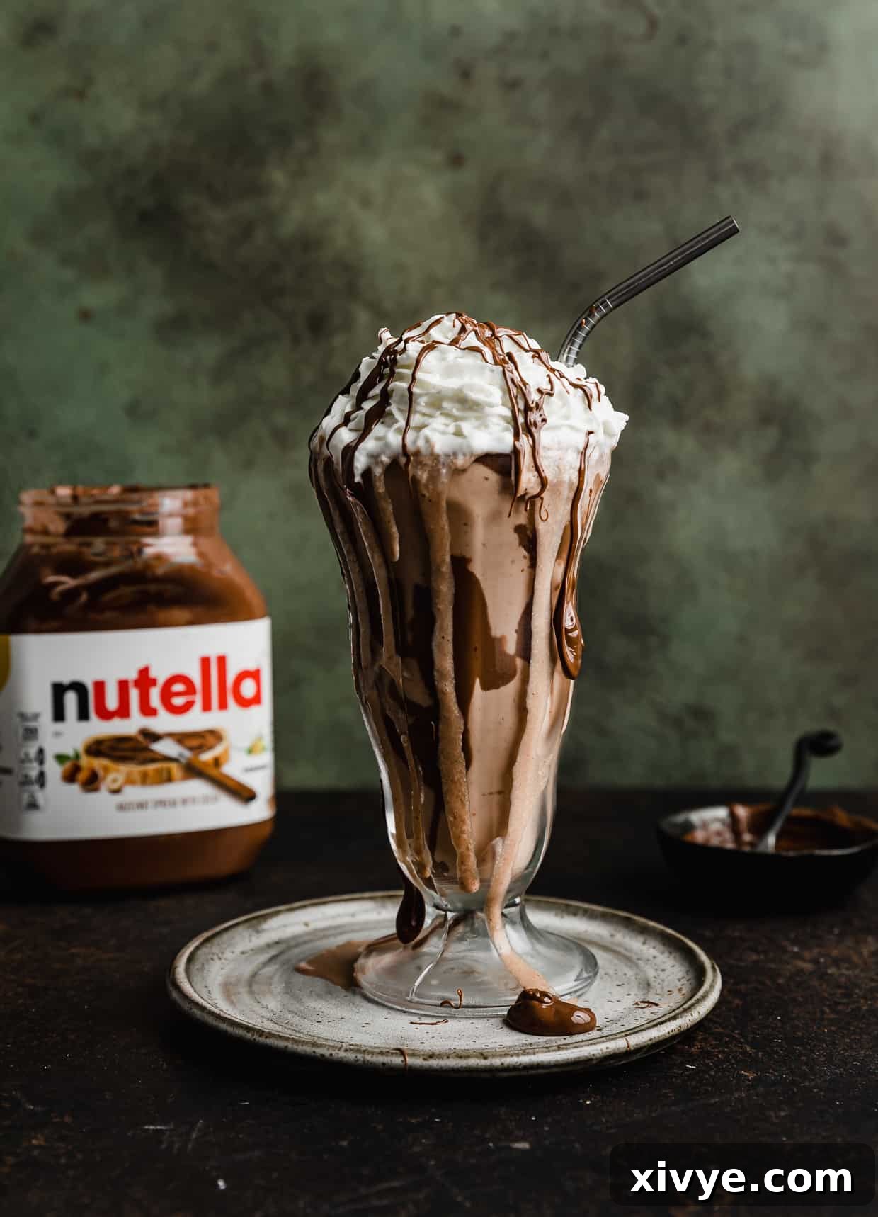 A tall glass milkshake cup filled with a rich Nutella Milkshake, generously topped with fluffy whipped cream and a luscious Nutella drizzle, set against a stylish dark green background.
