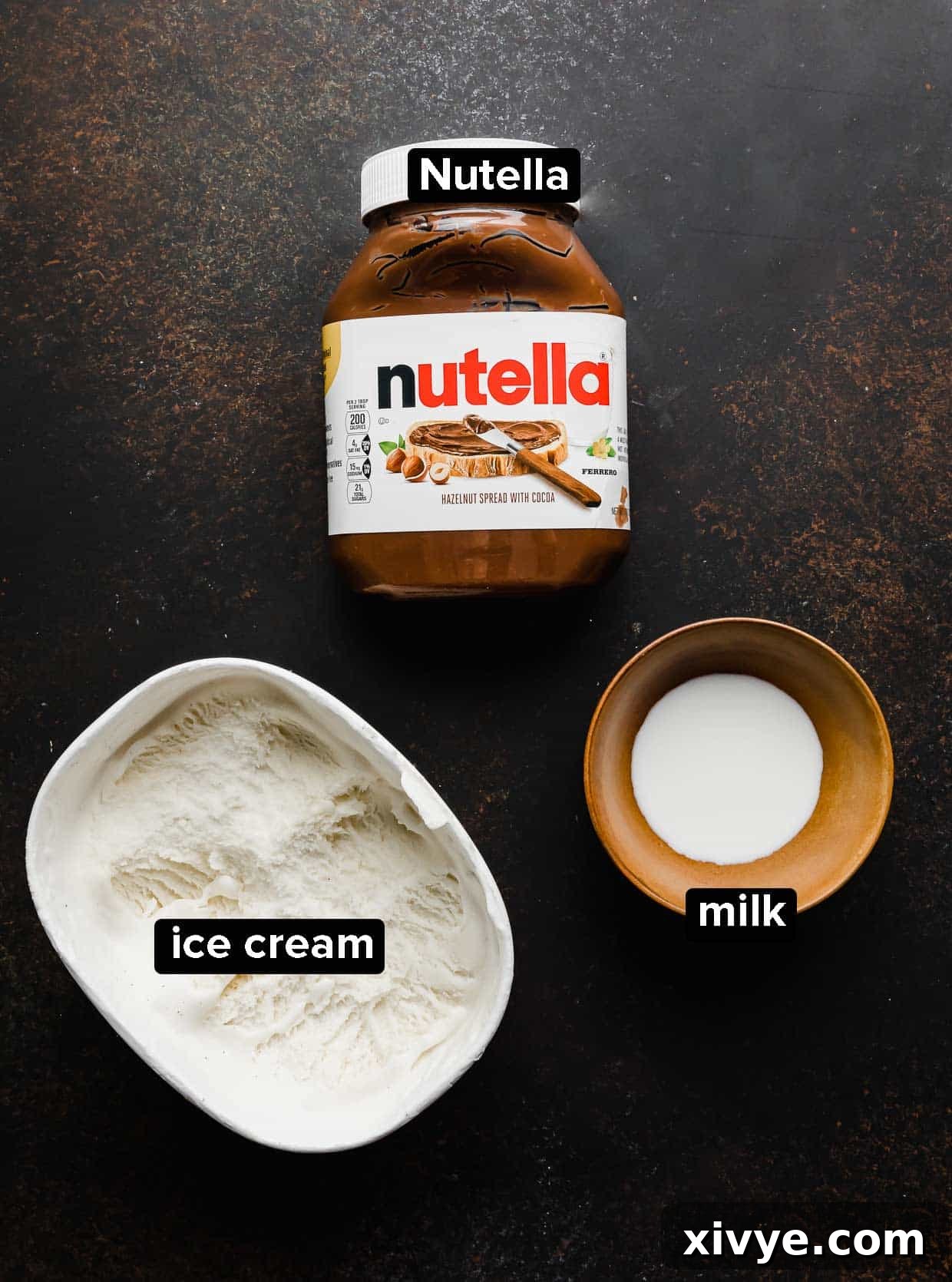 The simple yet powerful ingredients for a delicious Nutella Milkshake: a jar of Nutella, a tub of creamy vanilla ice cream, and a carton of milk, all arranged on a dark brown background.