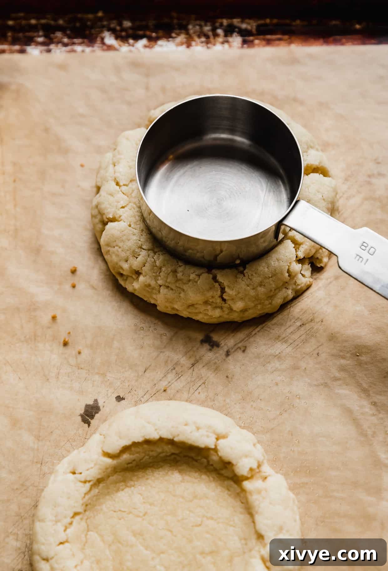 A measuring cup pressing into a partially baked sugar cookie to create an indentation.